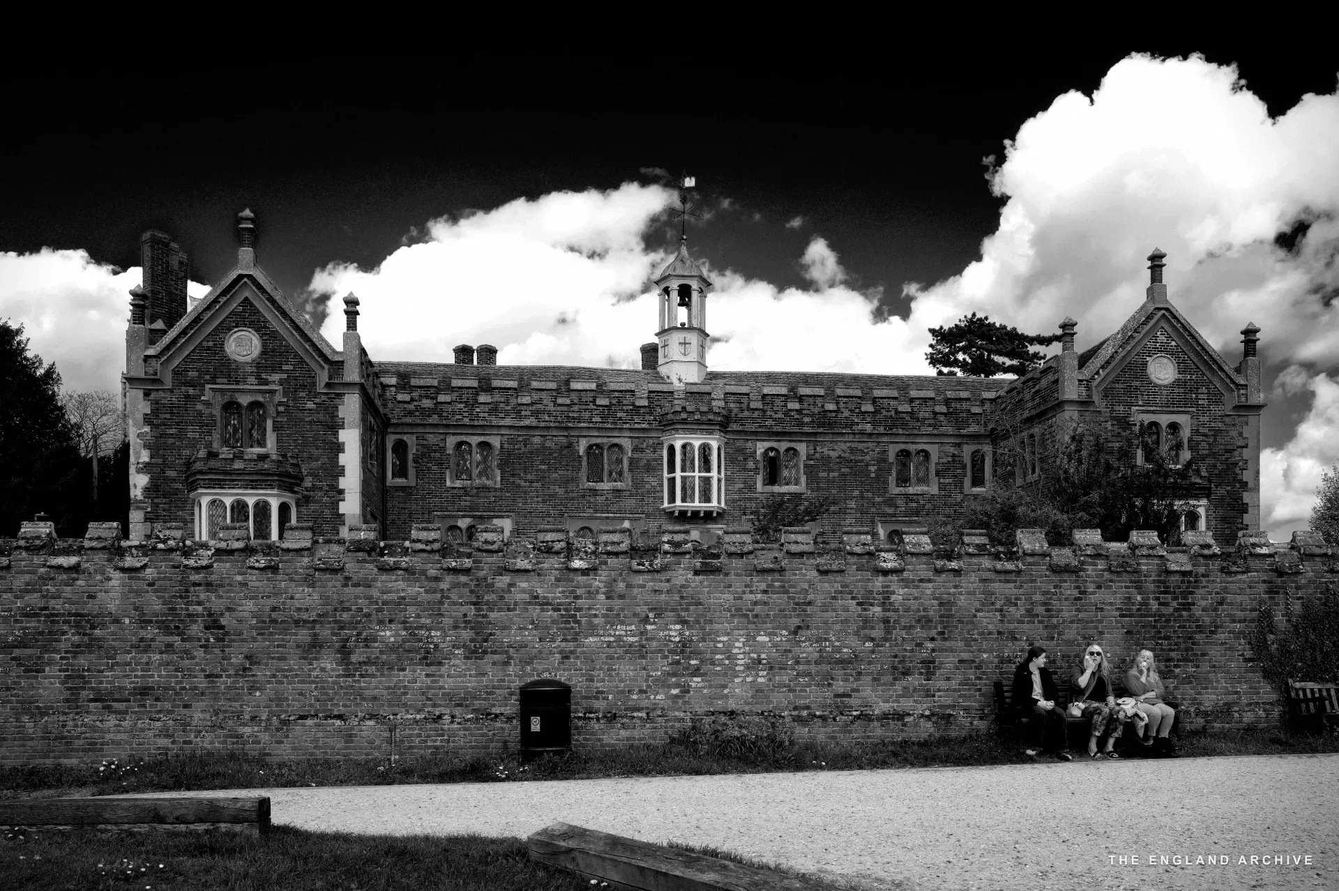 A long view of Trinity Hospital behind its raised red brick outer wall, central cupola above, three figures seated on a bench against the wall.