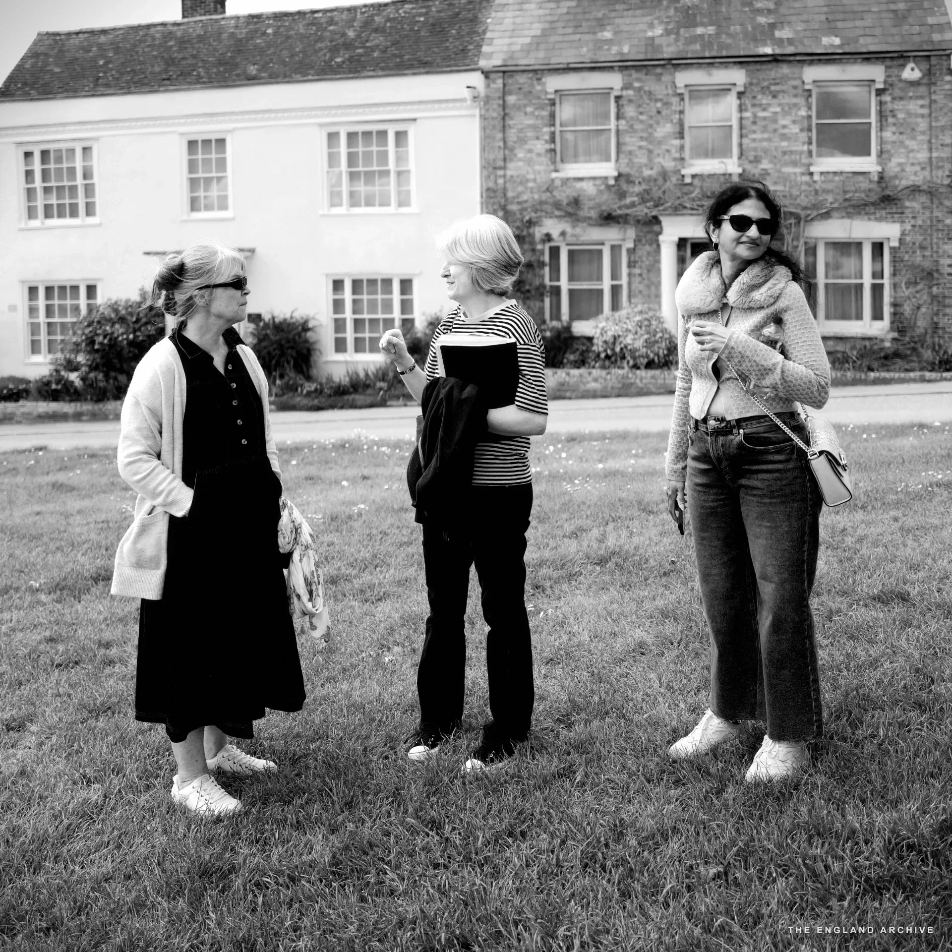 Julie Thomson, Melonie Clubb and Bhavani standing together on the grass of the Green, pale pink and cream Georgian houses behind them.