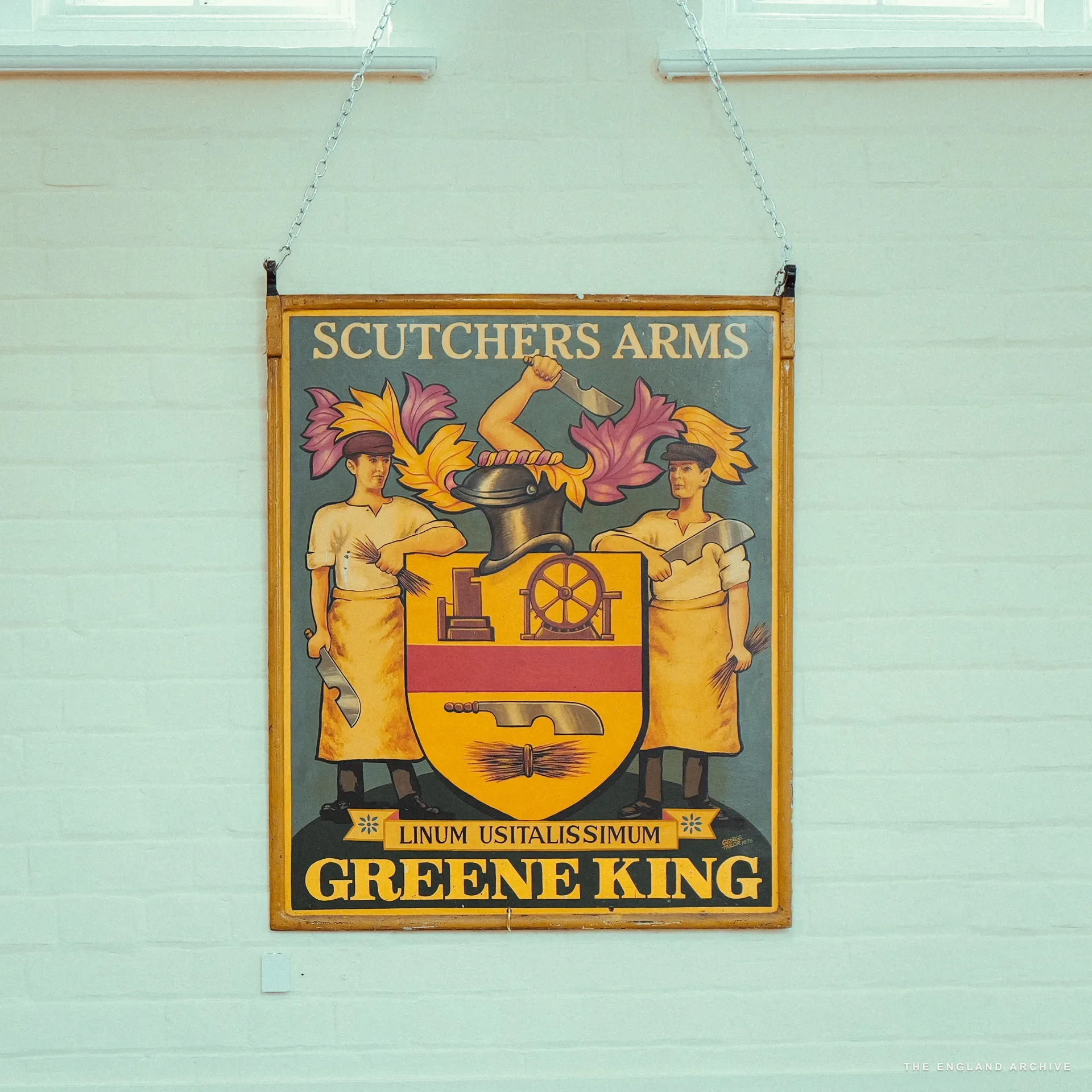 An old painted pub sign reading 'Scutchers Arms' showing a Greene King coat of arms with two flanking figures, hung against a pale mint-green wall.