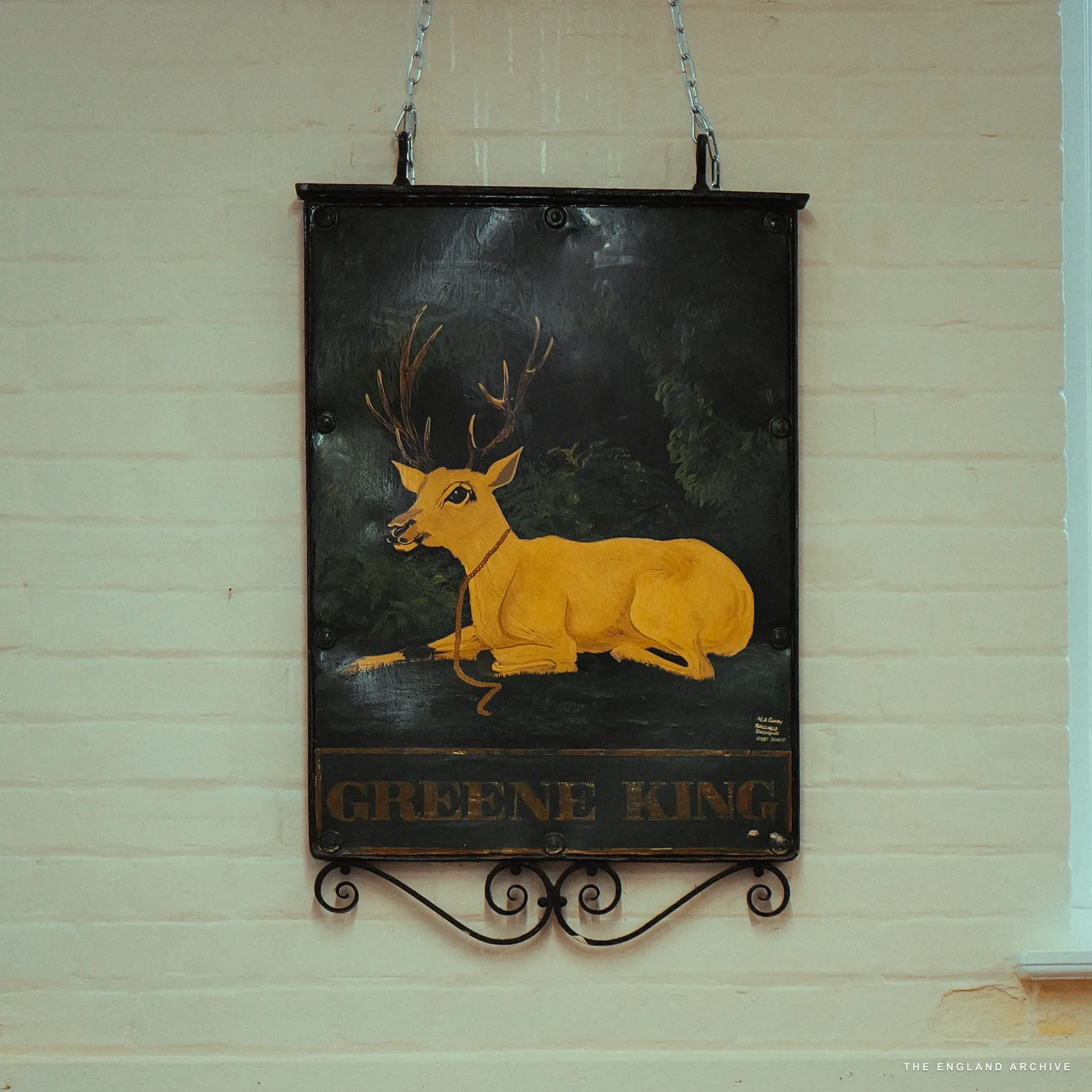 A Greene King pub sign with a painted yellow stag, hung on chains against a cream brick wall.