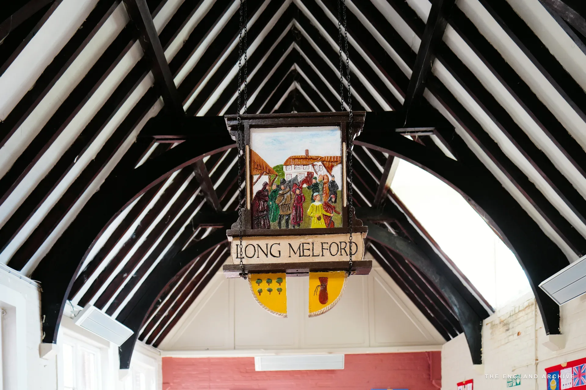 A carved wooden hanging sign reading 'Long Melford' with a painted village scene, beneath the steep timber-trussed roof of the community hall.