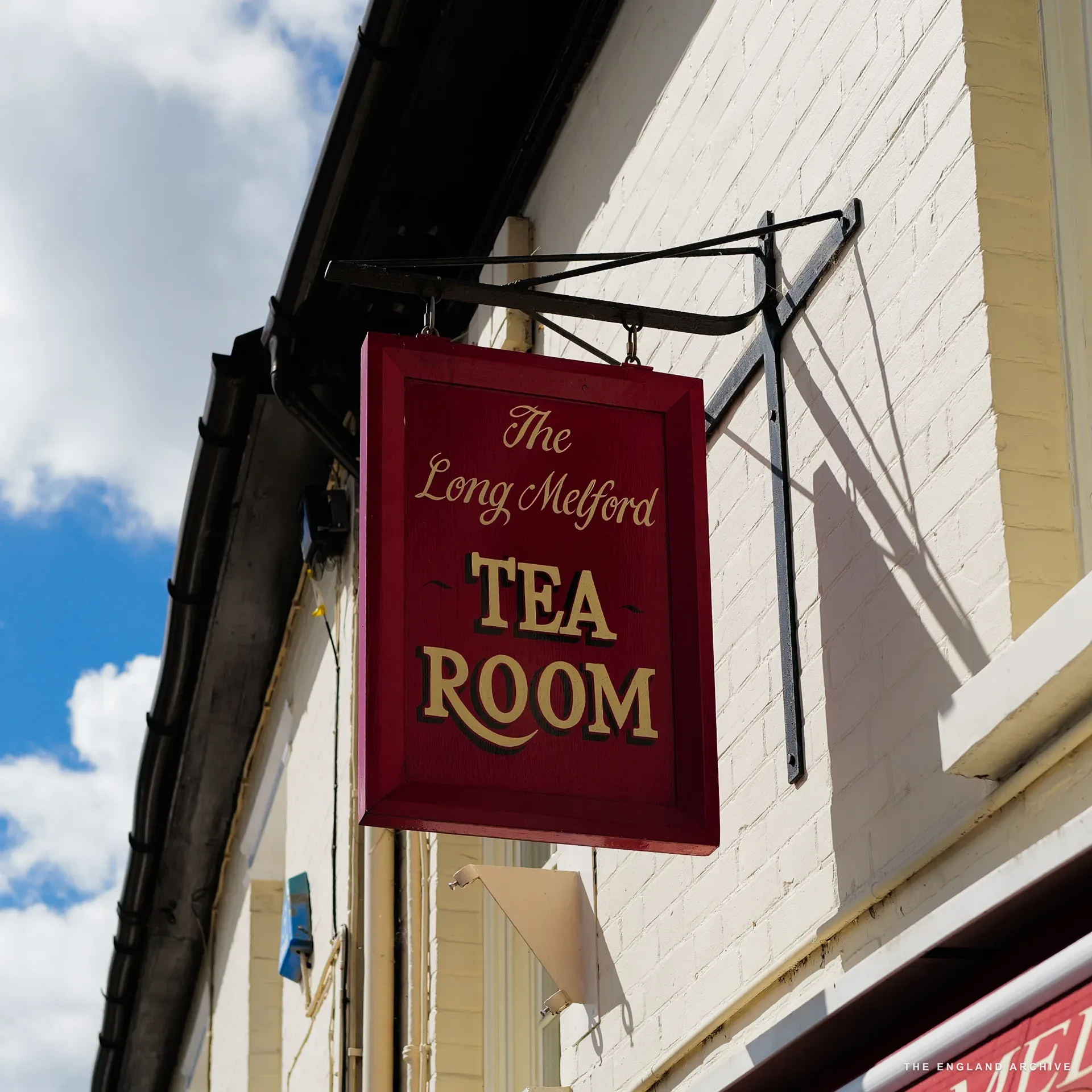A red and gold hanging sign reading 'The Long Melford TEA ROOM' with a decorative iron bracket against a white building.