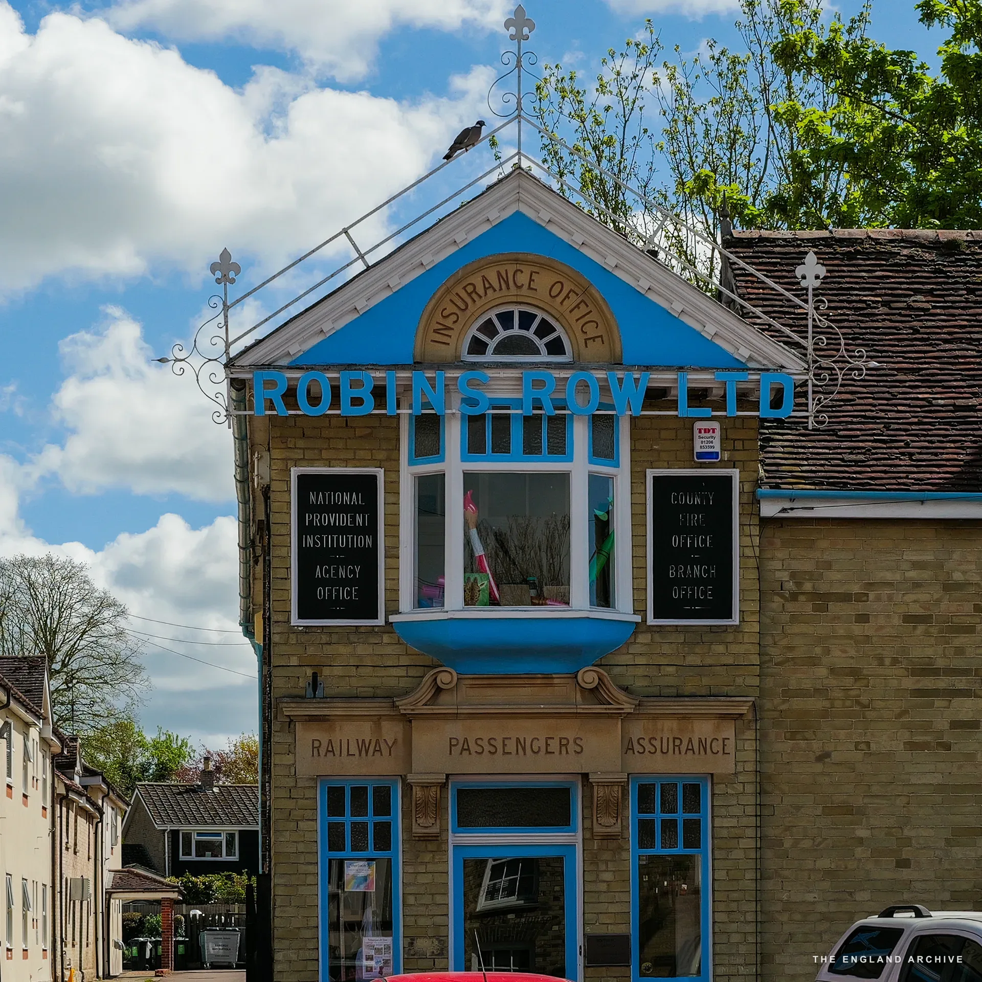 A quirky yellow-brick building with ornate blue signage reading 'Robins Row Ltd' and a 'RAILWAY PASSENGERS ASSURANCE' banner - a former insurance office preserved as its own period piece.