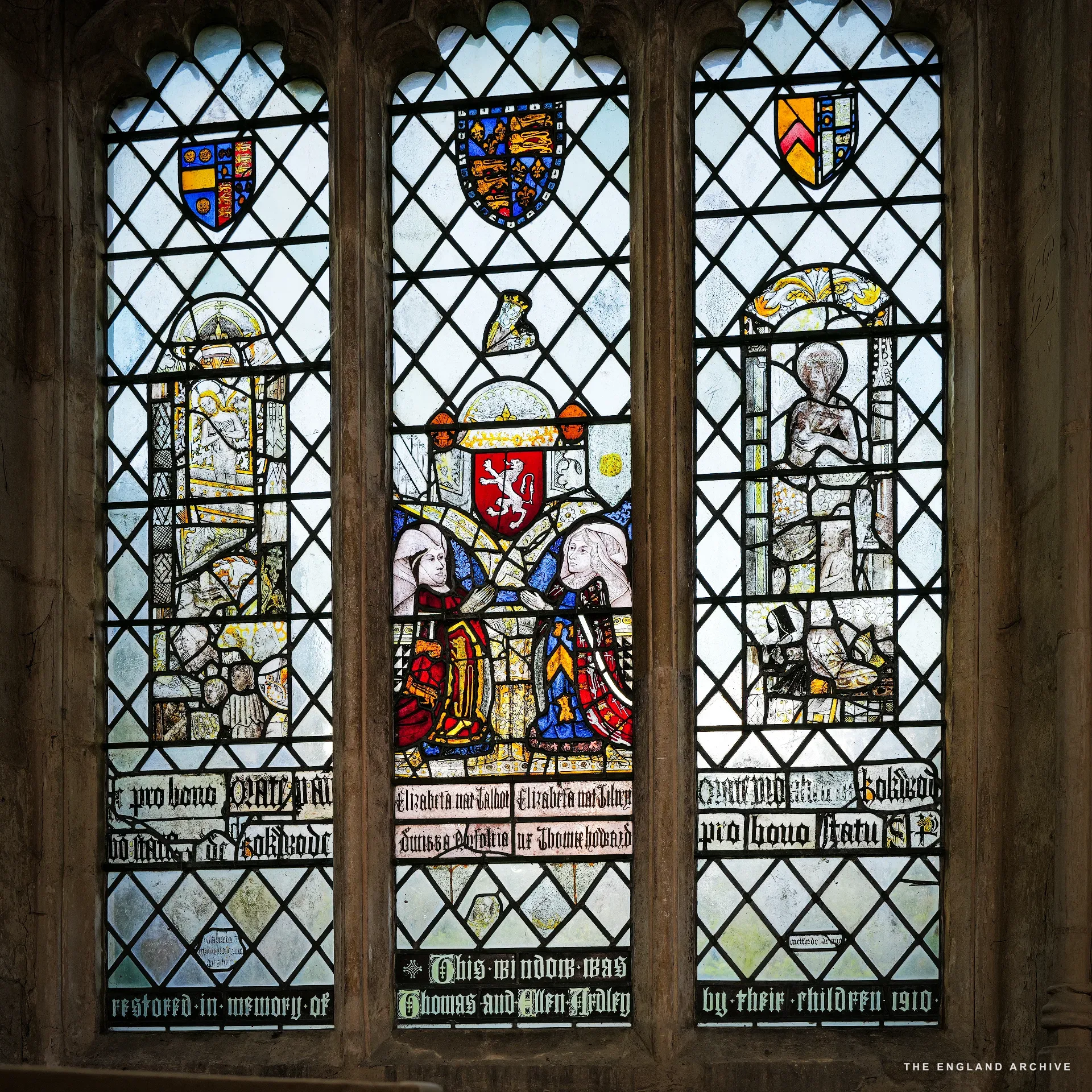 A three-light medieval stained-glass window with kneeling donor figures - a 1910 memorial to Thomas and Ellen Ardley - with Latin inscription below.
