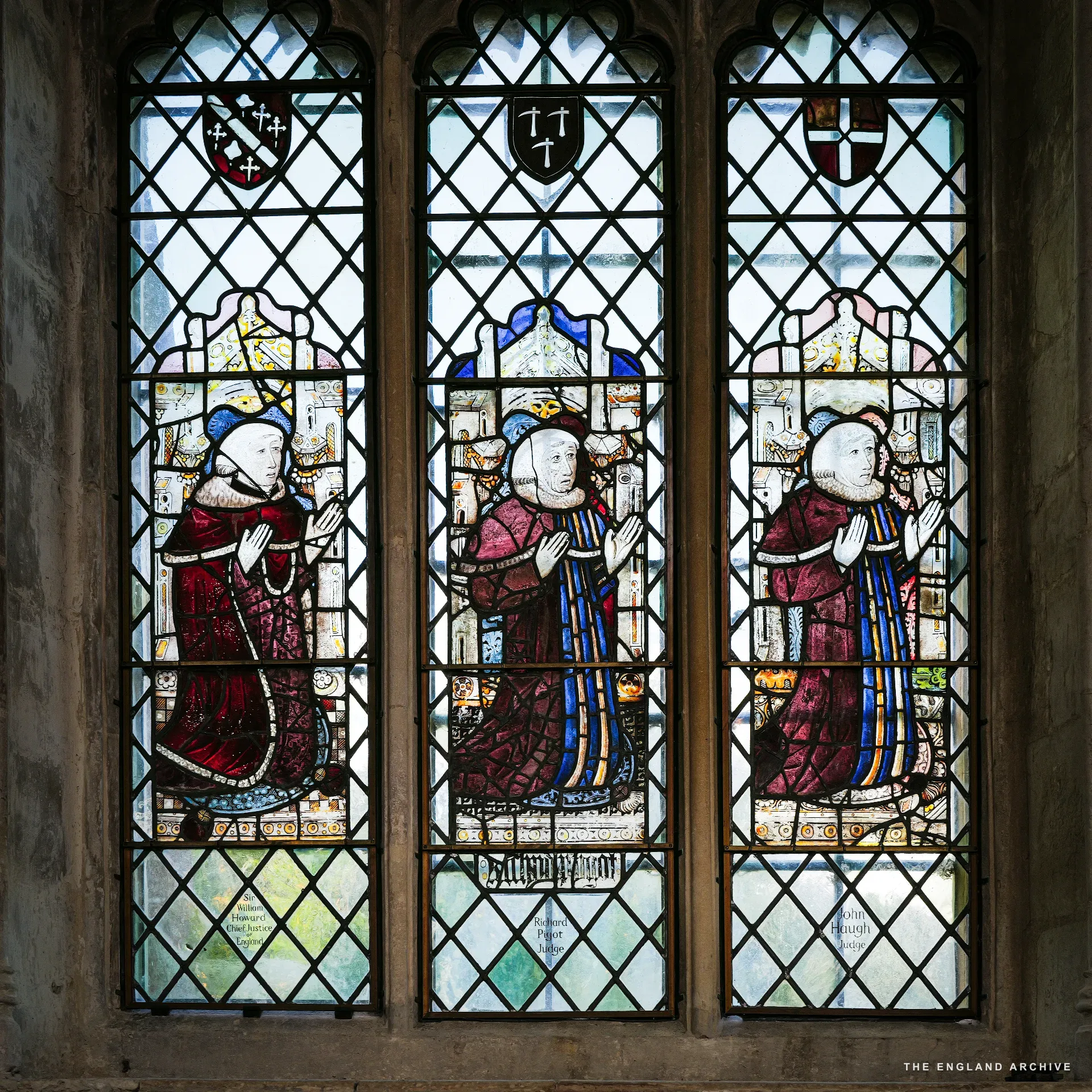 A medieval stained-glass three-light window of kneeling ecclesiastical figures in red and blue robes.