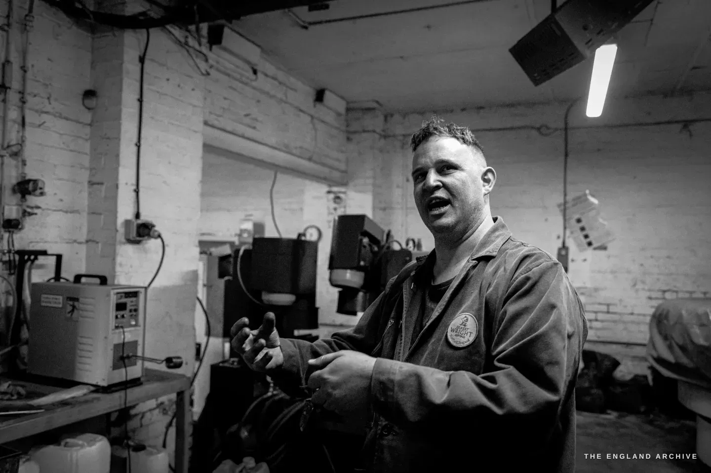 Neil mid-sentence on the workshop floor, hands raised in explanation, the brick room and machinery behind him.