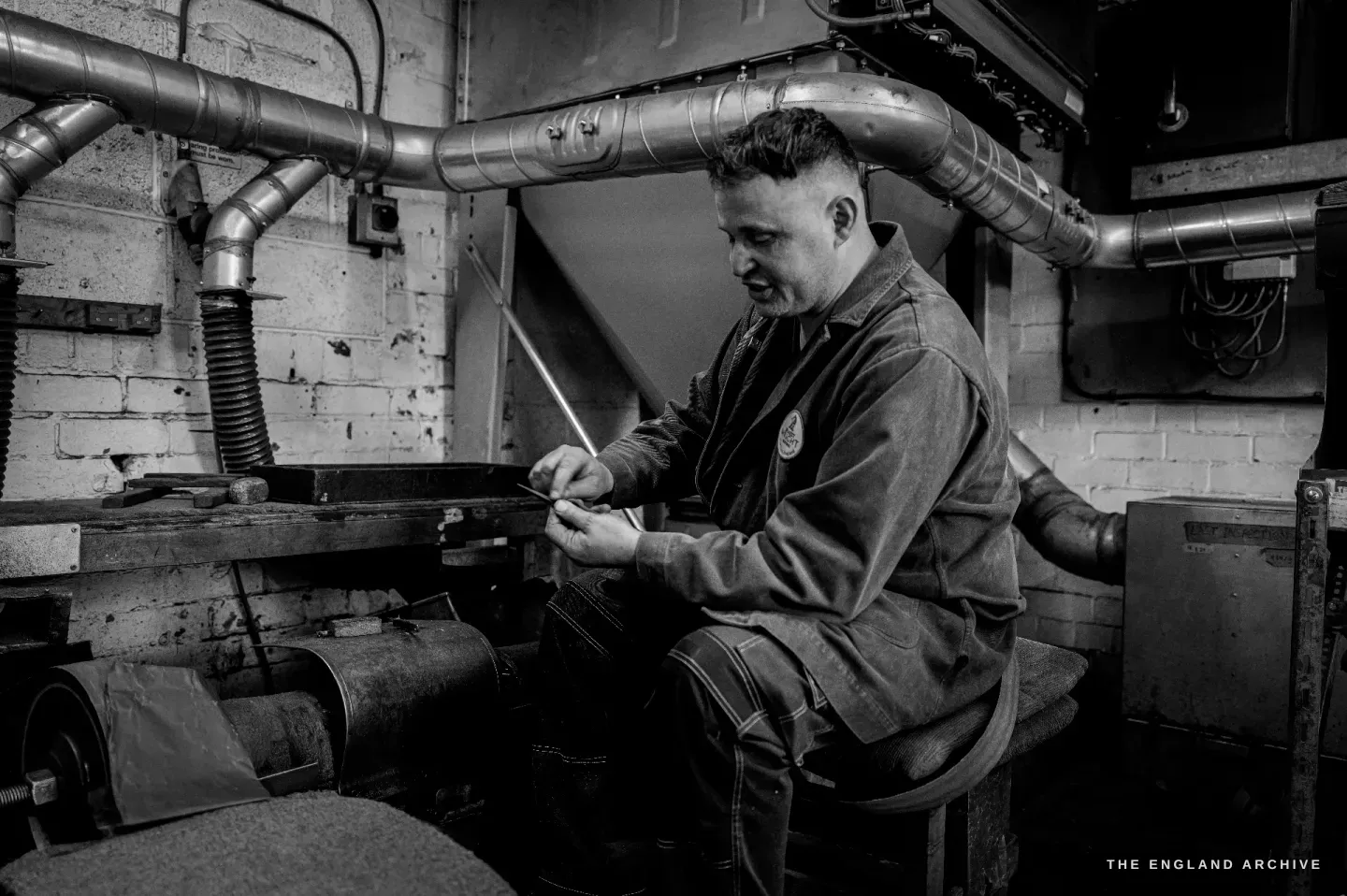 Neil seated at a bench beneath the extraction ducting, working a long blade in his hands, racks of grindstones beside him.