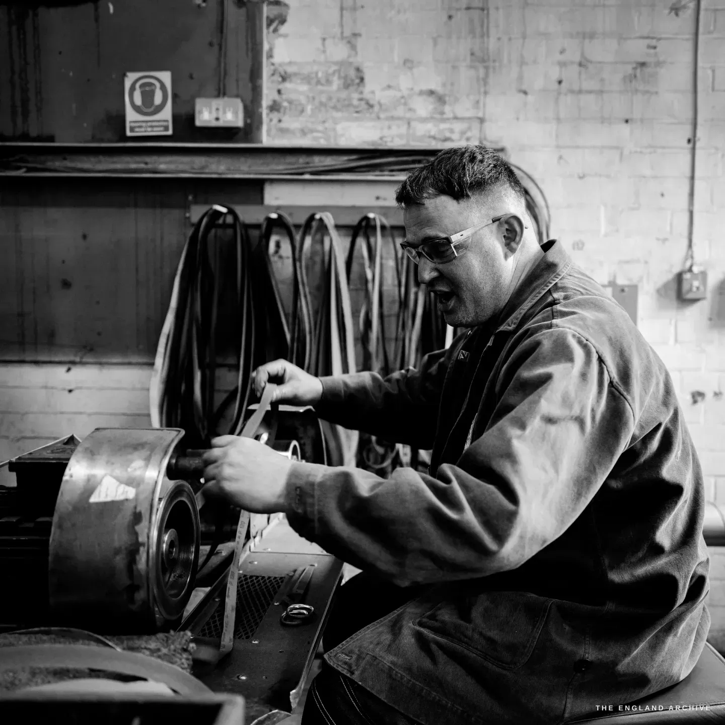 Neil leaning into a grinding wheel, glasses on, both hands guiding a blade, rows of leather drive belts on the wall behind.