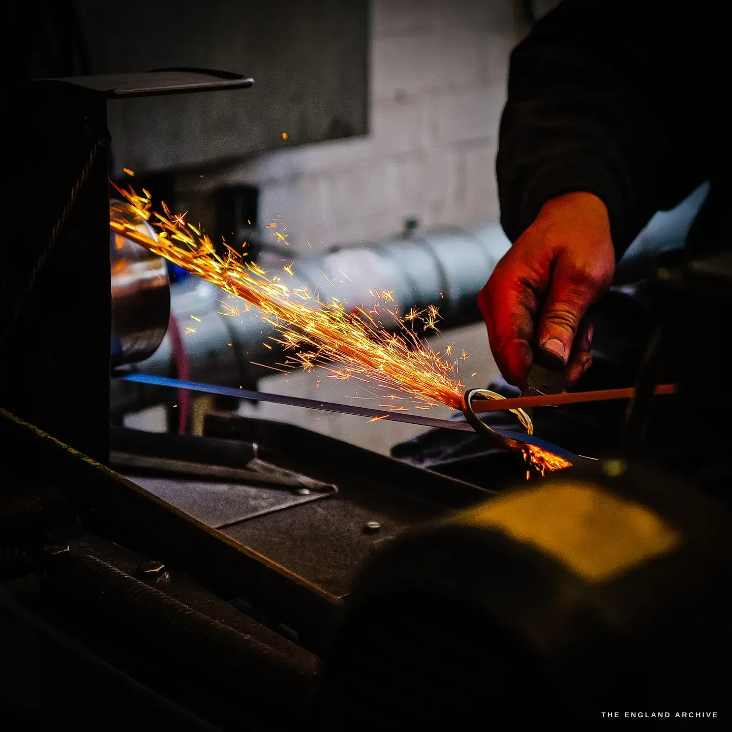 A close low-key frame of Neil’s hand holding a blade to a wheel, a bright fan of sparks streaming off into the dark.