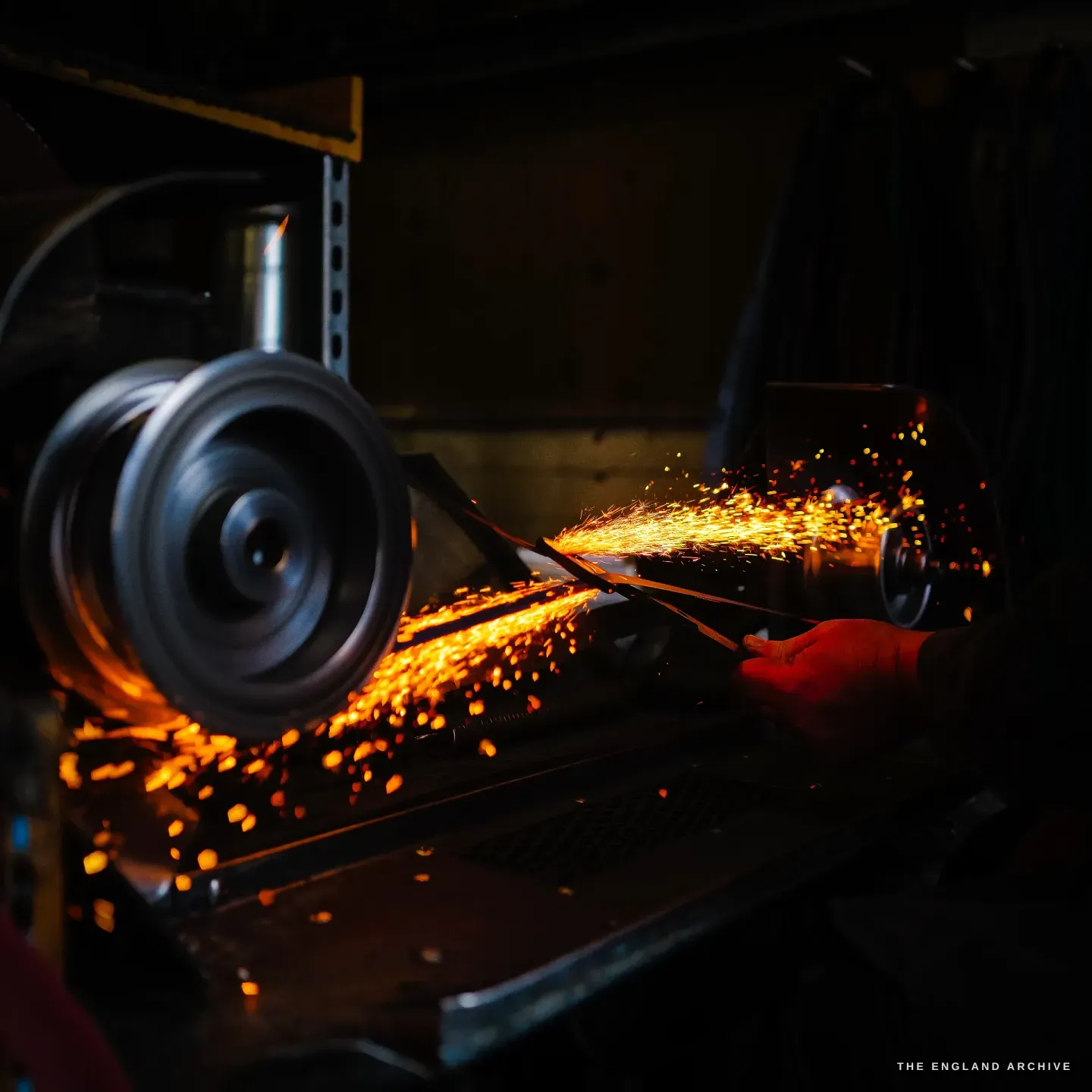 A blade held against a spinning wheel, sparks arcing across the frame, the leather belt and pulley lit behind.