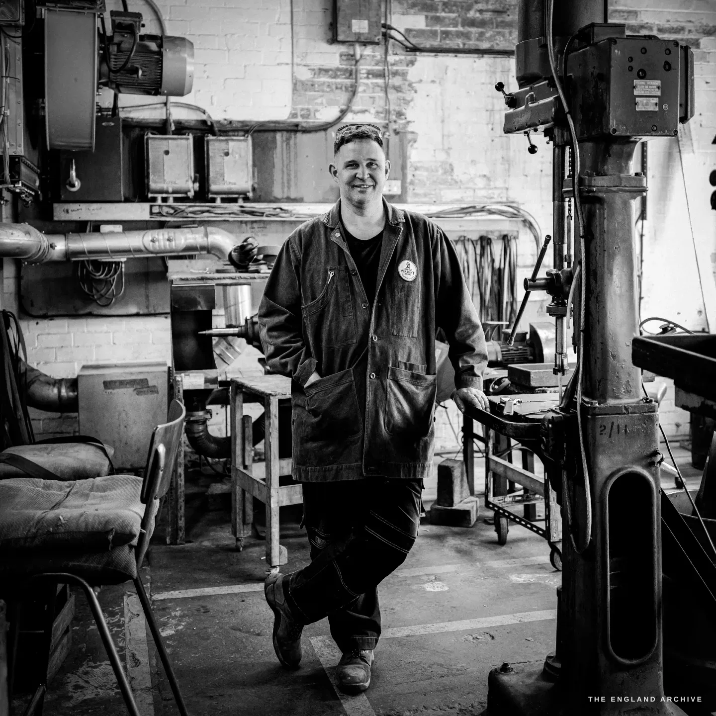 A full-length portrait of Neil leaning against a pillar drill in the workshop, relaxed and smiling, the long brick room stretching behind him.