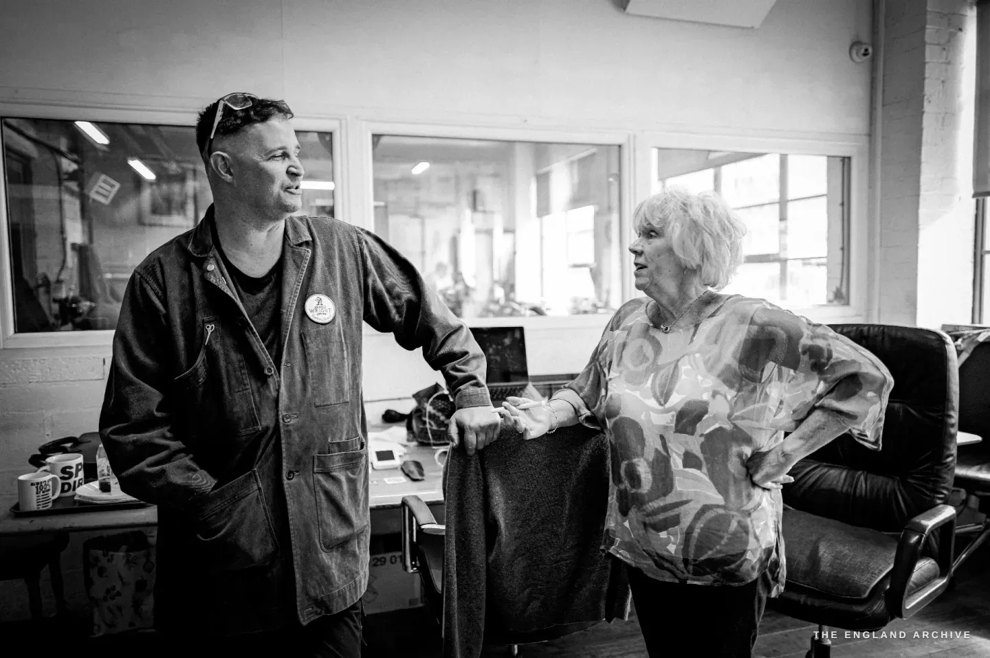 Neil in conversation with a colleague in the office area of the workshop, both smiling, windows and desks behind.