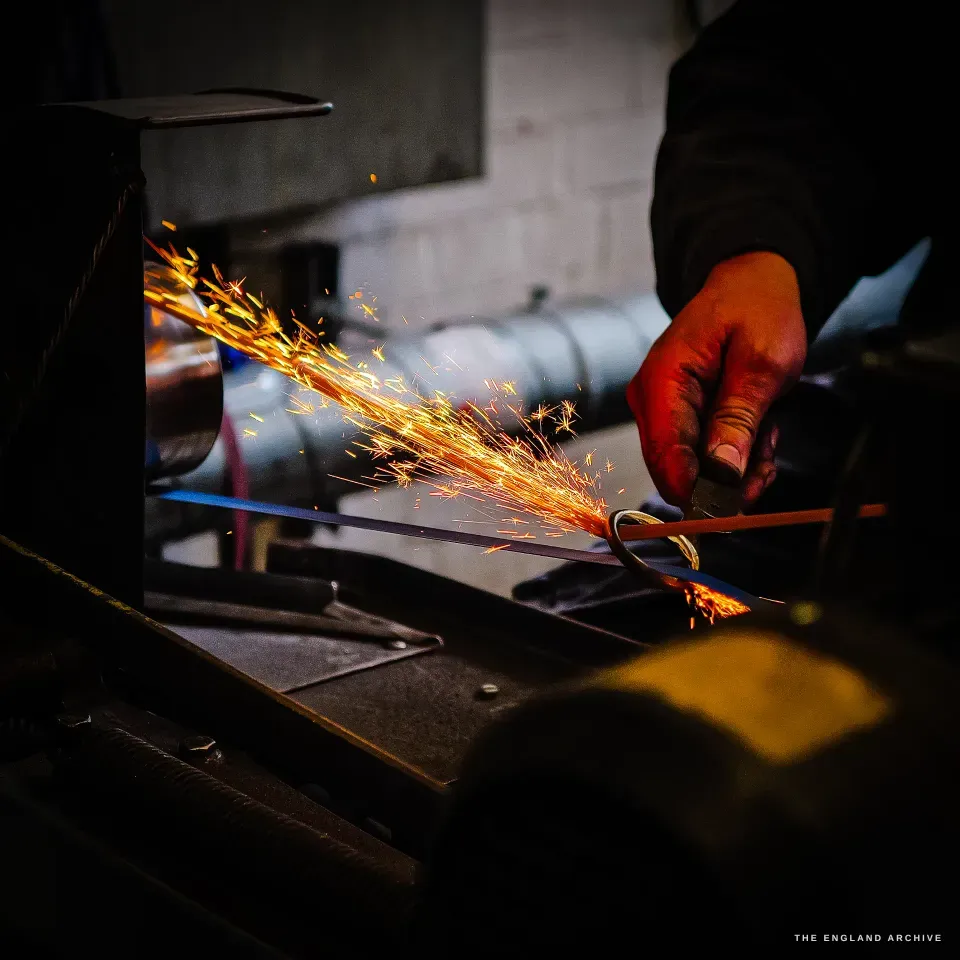 A close low-key frame of Neil’s hand holding a blade to a wheel, a bright fan of sparks streaming off into the dark.