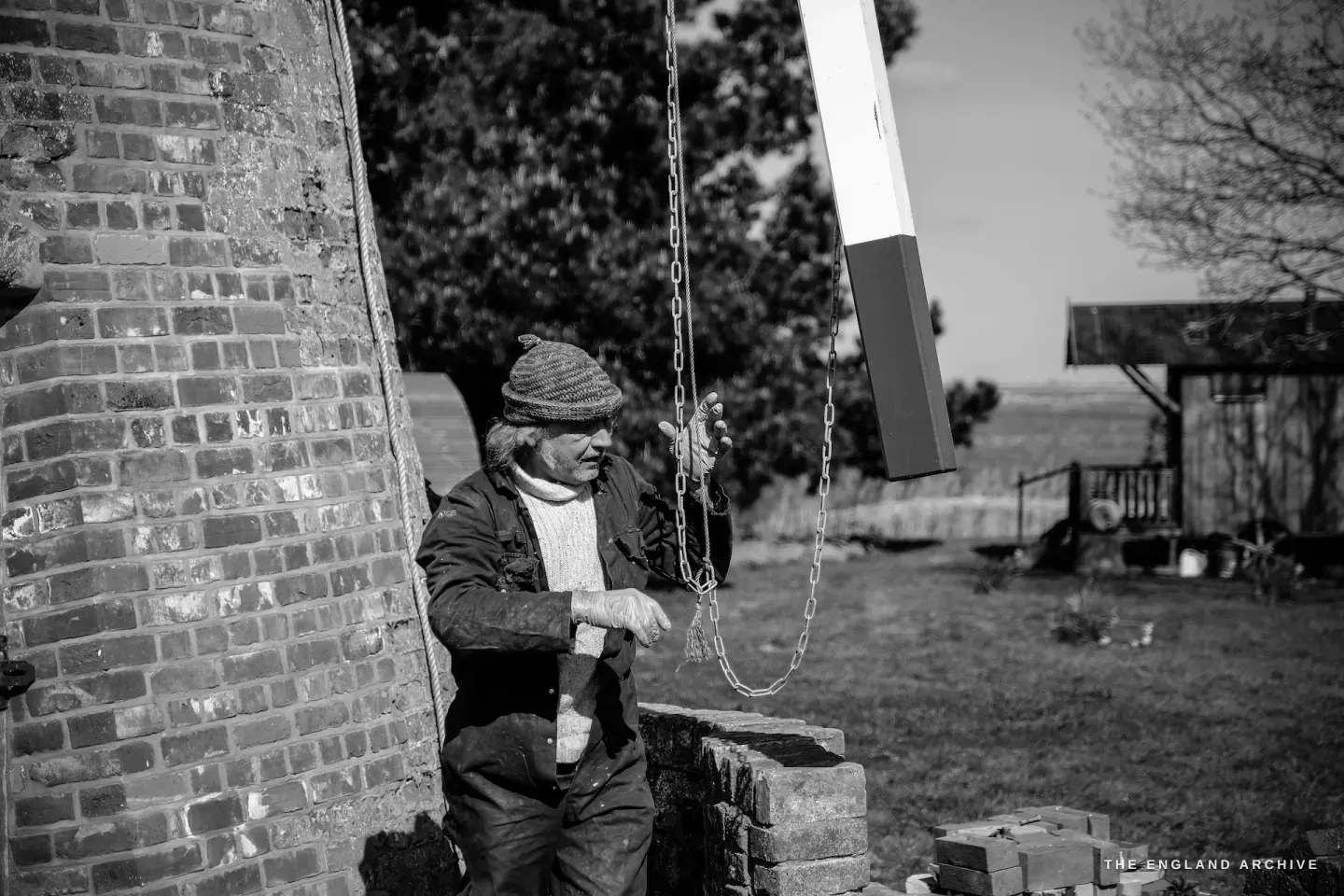 Paul Kemp leaning against the mill brickwork, one hand on the chain, looking out across the marshes