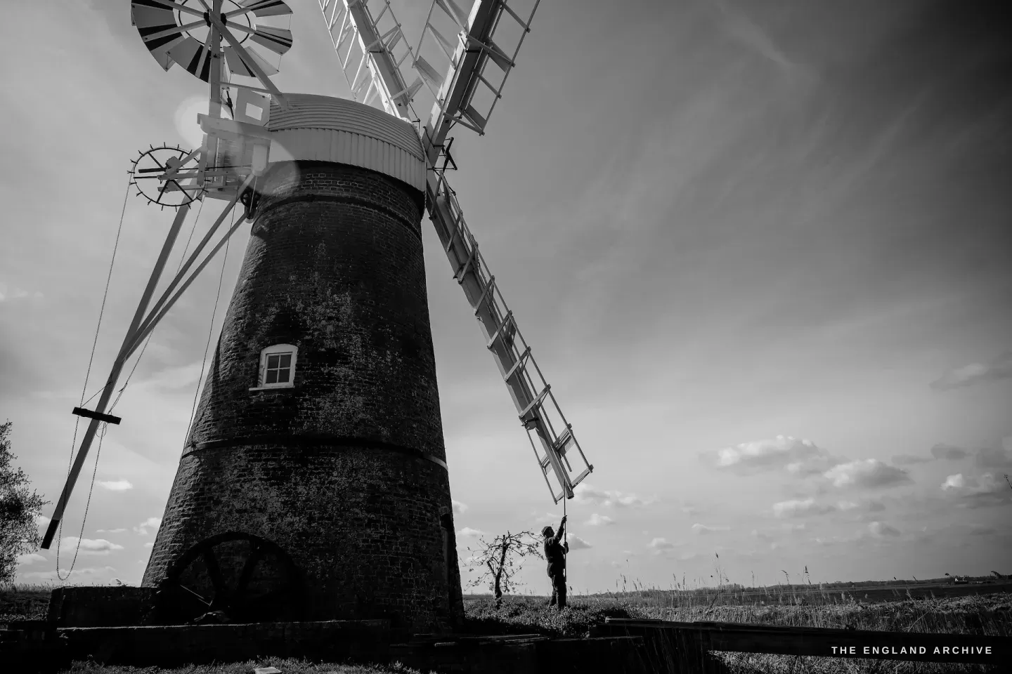A dramatic wide-angle view of the full mill from below, Paul visible beside the base, the sails towering above against cloud