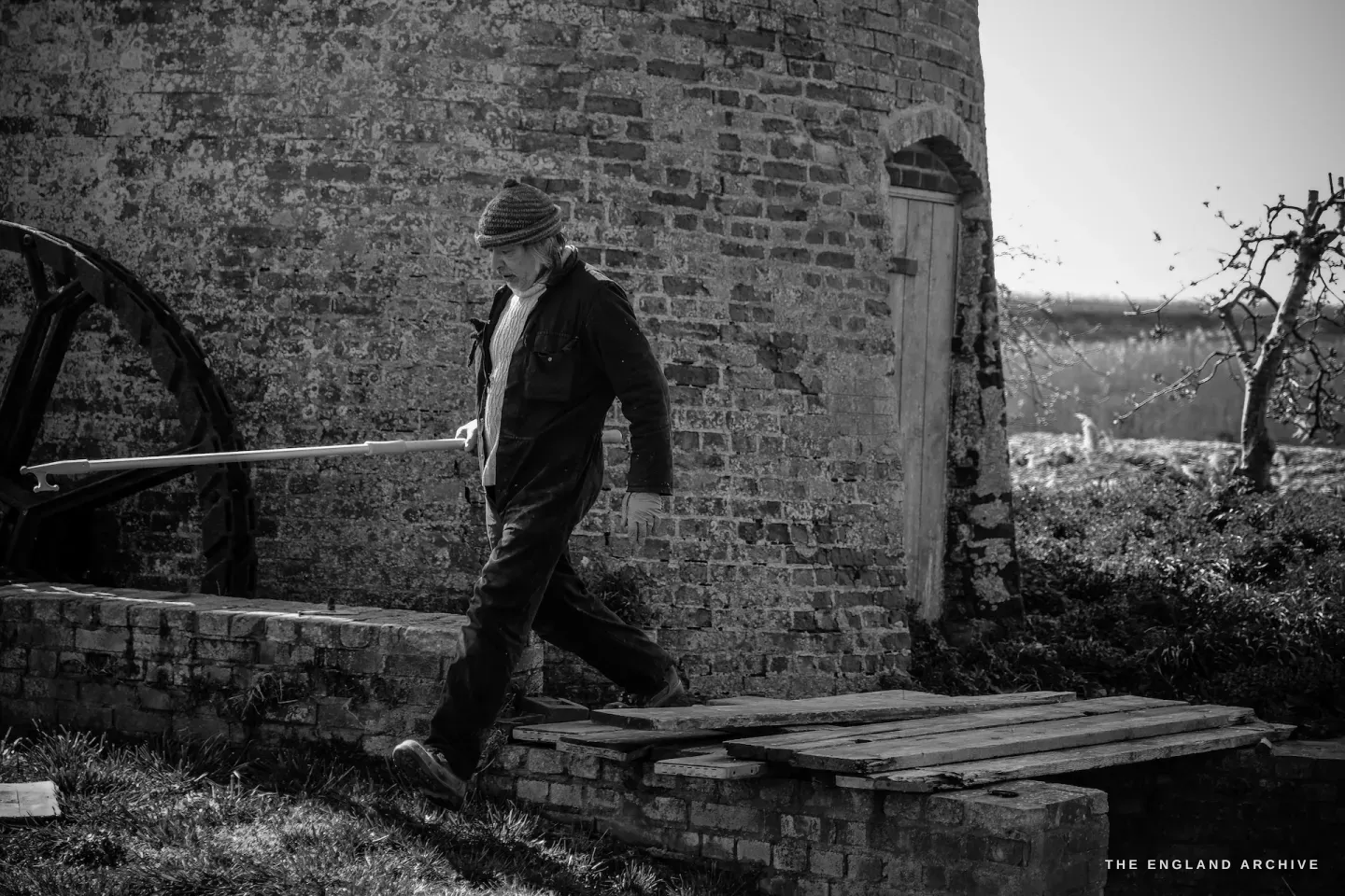Paul Kemp walking past the mill base, brickwork and a wooden pallet beside him, moving with unhurried purpose