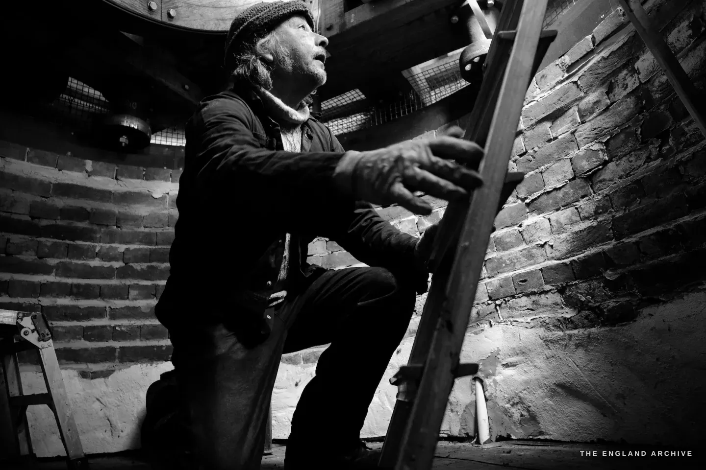 Paul on the interior ladder of the mill, looking upward, dramatic low light from the window illuminating his face and the brickwork