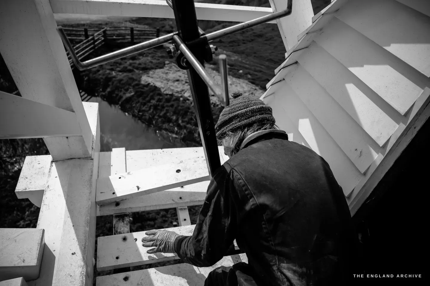 Paul climbing down through the exterior sail framework, seen from above, the ground and marshland below