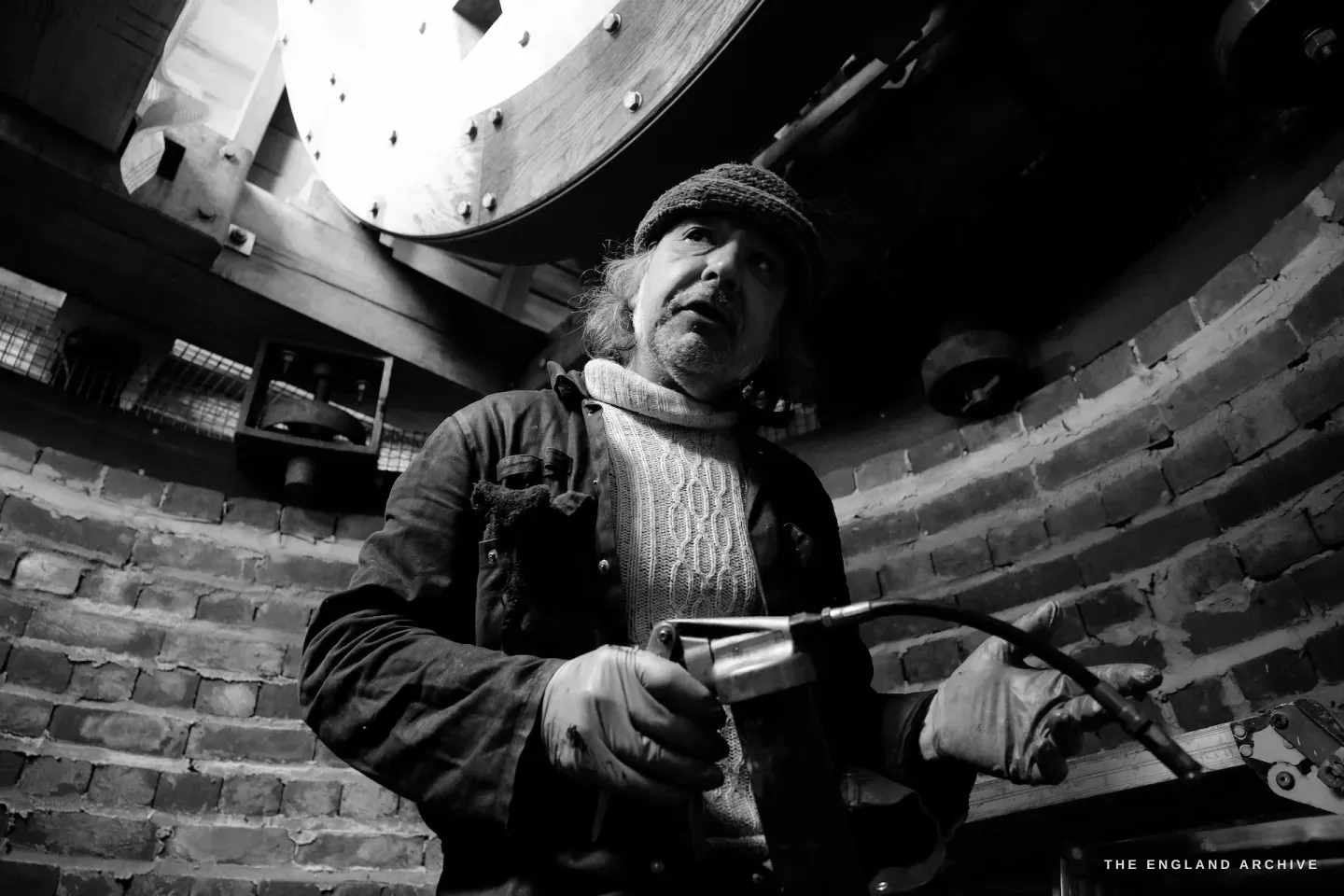 Paul holding a grease gun inside the mill, the large wooden gear wheel behind him