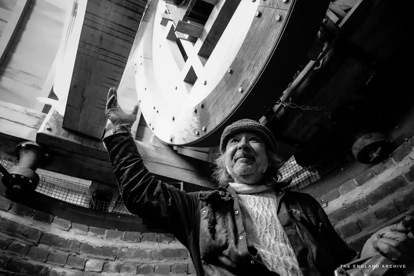 Paul reaching up to touch the large wooden gear wheel inside the mill, looking up at the mechanism with familiarity