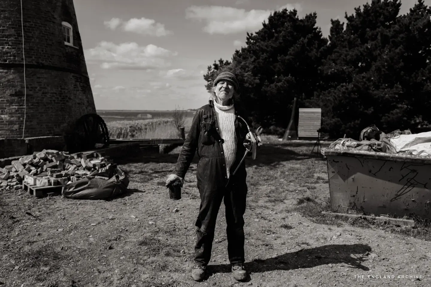 Paul standing full-length at the mill, holding a grease gun, looking at the camera, the tower and piled materials behind him