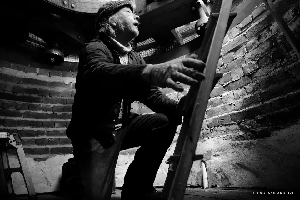 Paul on the interior ladder of the mill, looking upward, dramatic low light from the window illuminating his face and the brickwork