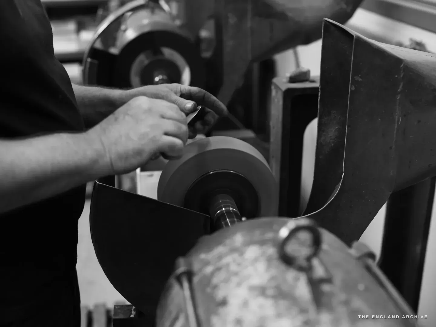Over Paul’s shoulder, both hands hold a small blade to a tapered polishing wheel on a spindle, a second wheel turning behind, a galvanised collecting drum below.