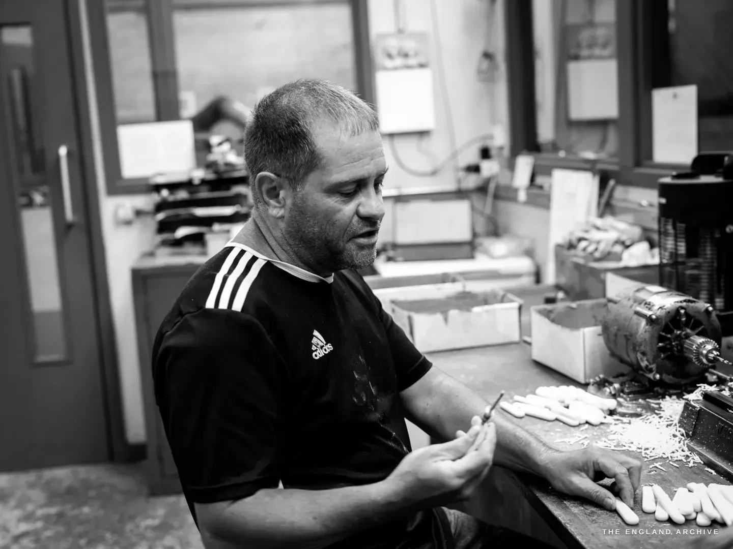 Paul Weatherstone seated at his bench, turning a knife in his hands beside a tray of pale cream handle blanks, drilling machinery to his right.