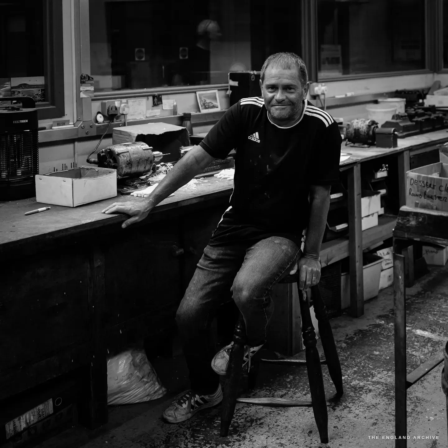Paul Weatherstone perched on a stool against his workbench, arms easy, looking to camera with a half-smile, the finishing room behind him.