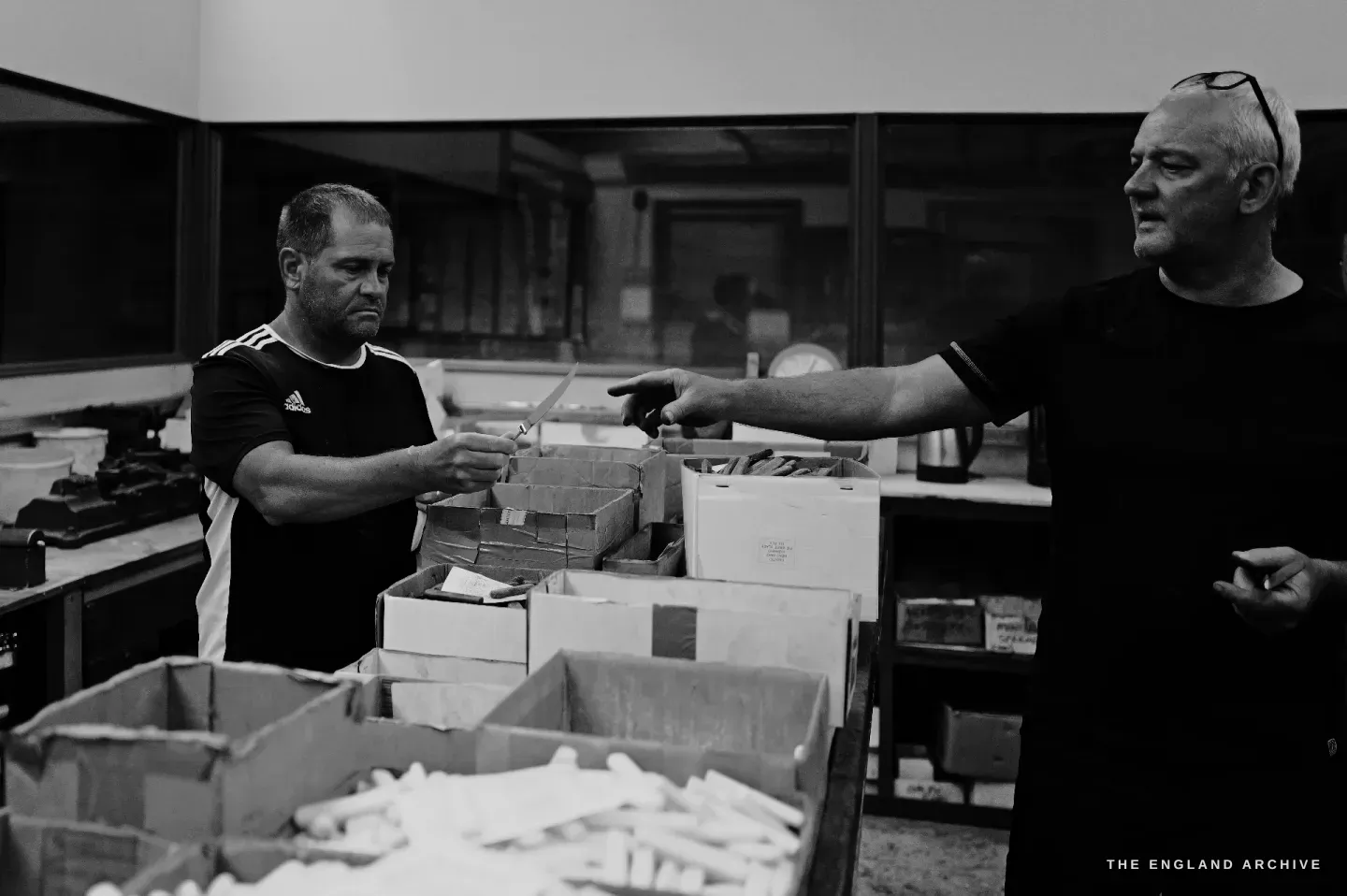 Phil Benton at right gestures across boxes of cut handle blanks toward Paul at left, the two men working between the sections.