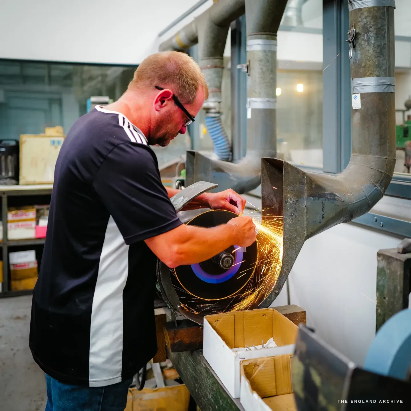 Paul in profile at a grinding wheel, a blade held to the rim, a fan of sparks thrown off into a cardboard box below.