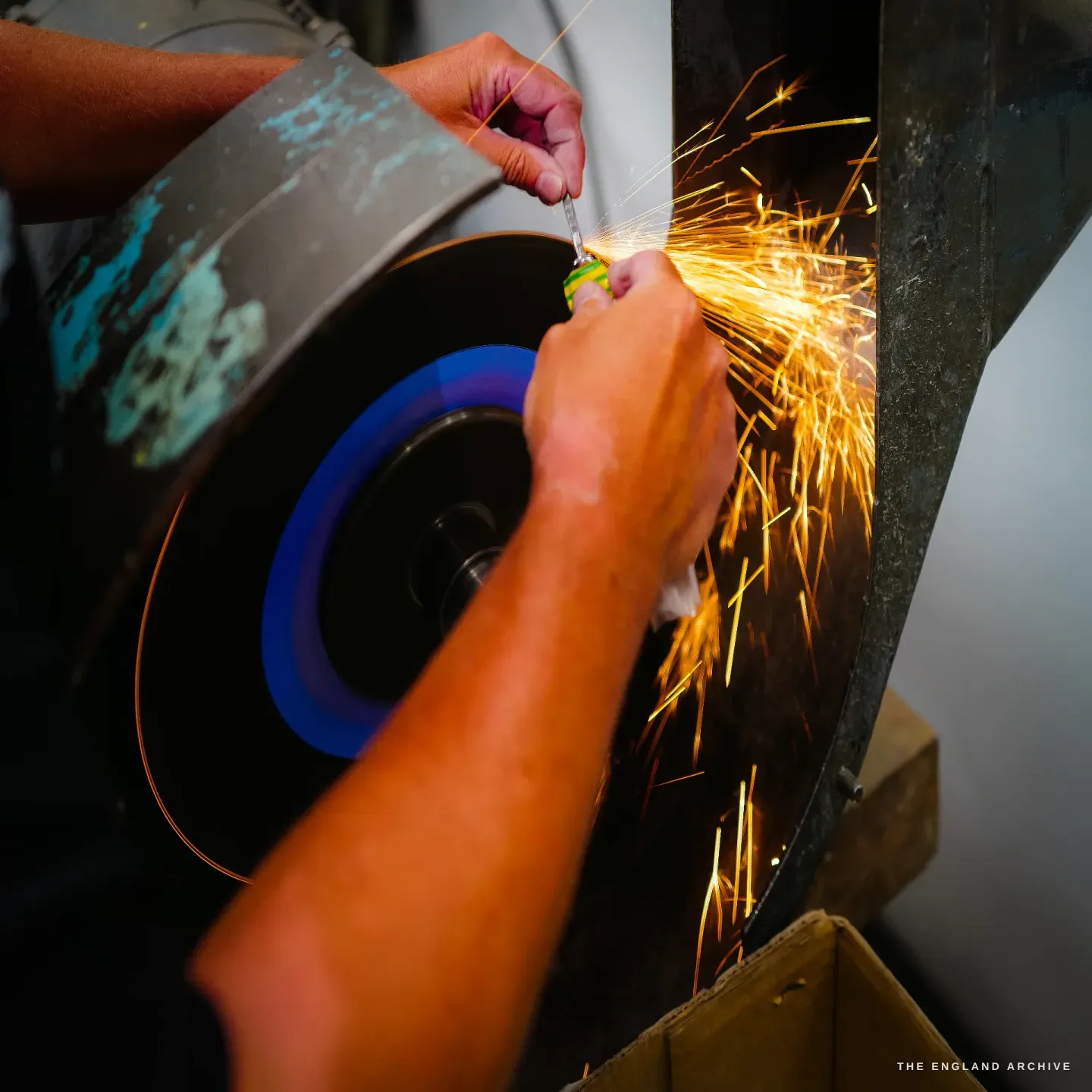 A close view of two hands holding a small knife against a spinning wheel, a bright spray of sparks arcing off to the side.