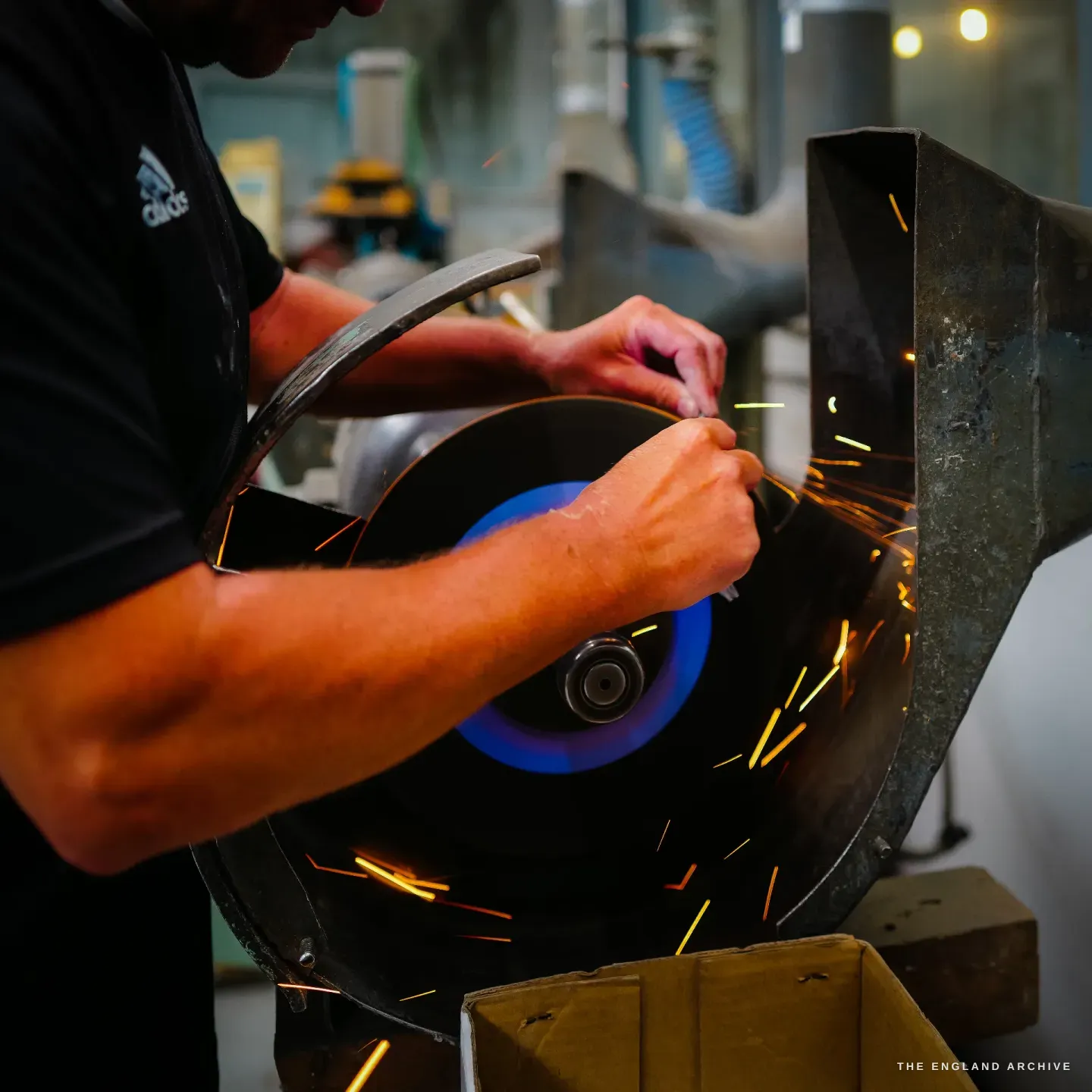 Paul leaning into a grinding wheel, both hands guiding a blade, sparks scattering from the contact point.