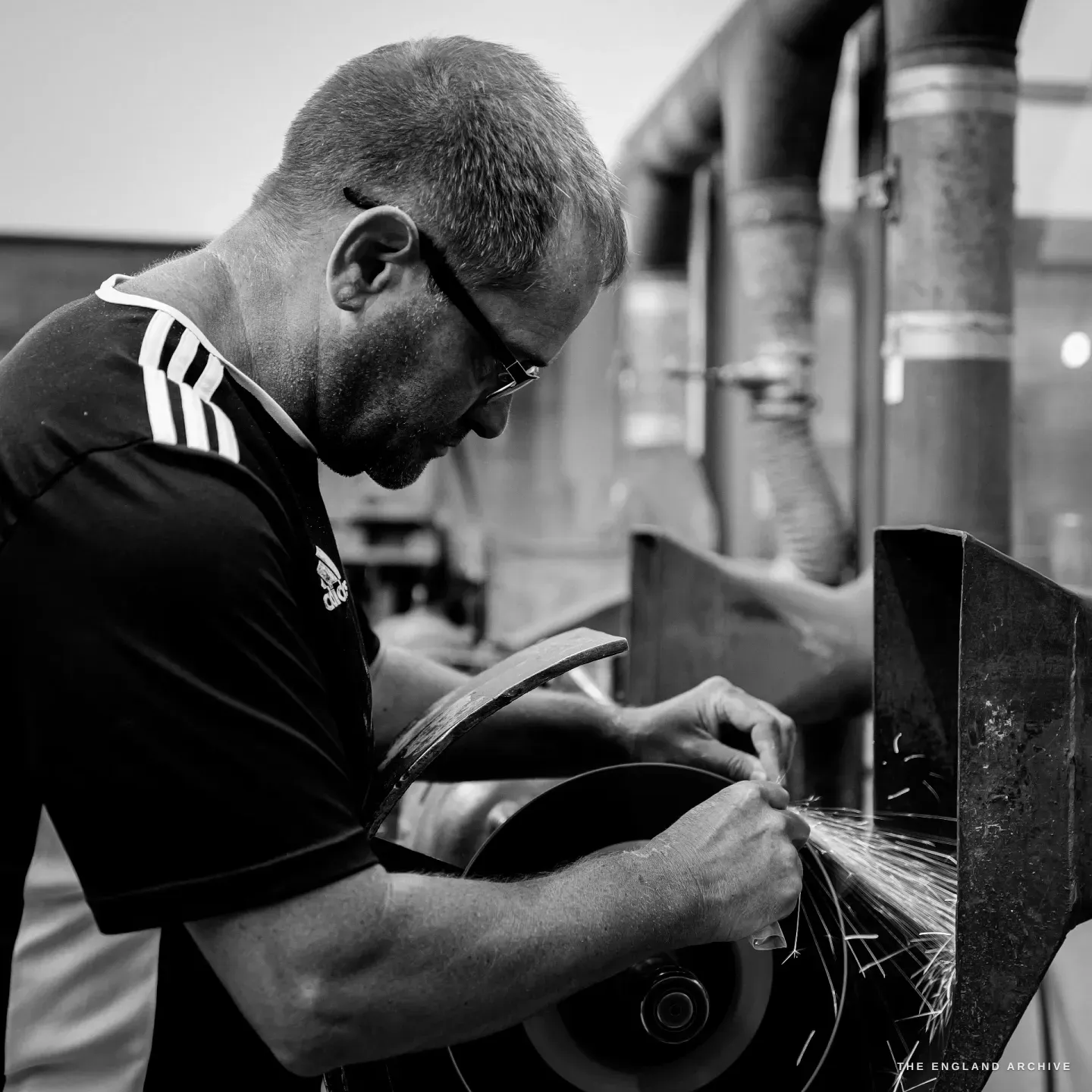 Paul in profile, glasses on, bent close over a grinding wheel as he edges a blade, fine sparks at the rim.