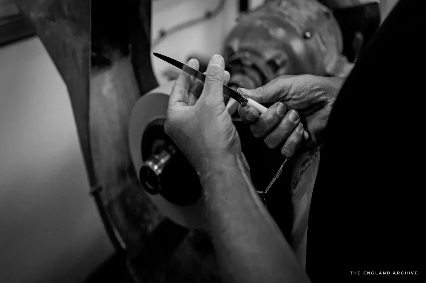 A low-key close-up of two weathered hands holding a slim pointed blade to a polishing wheel.