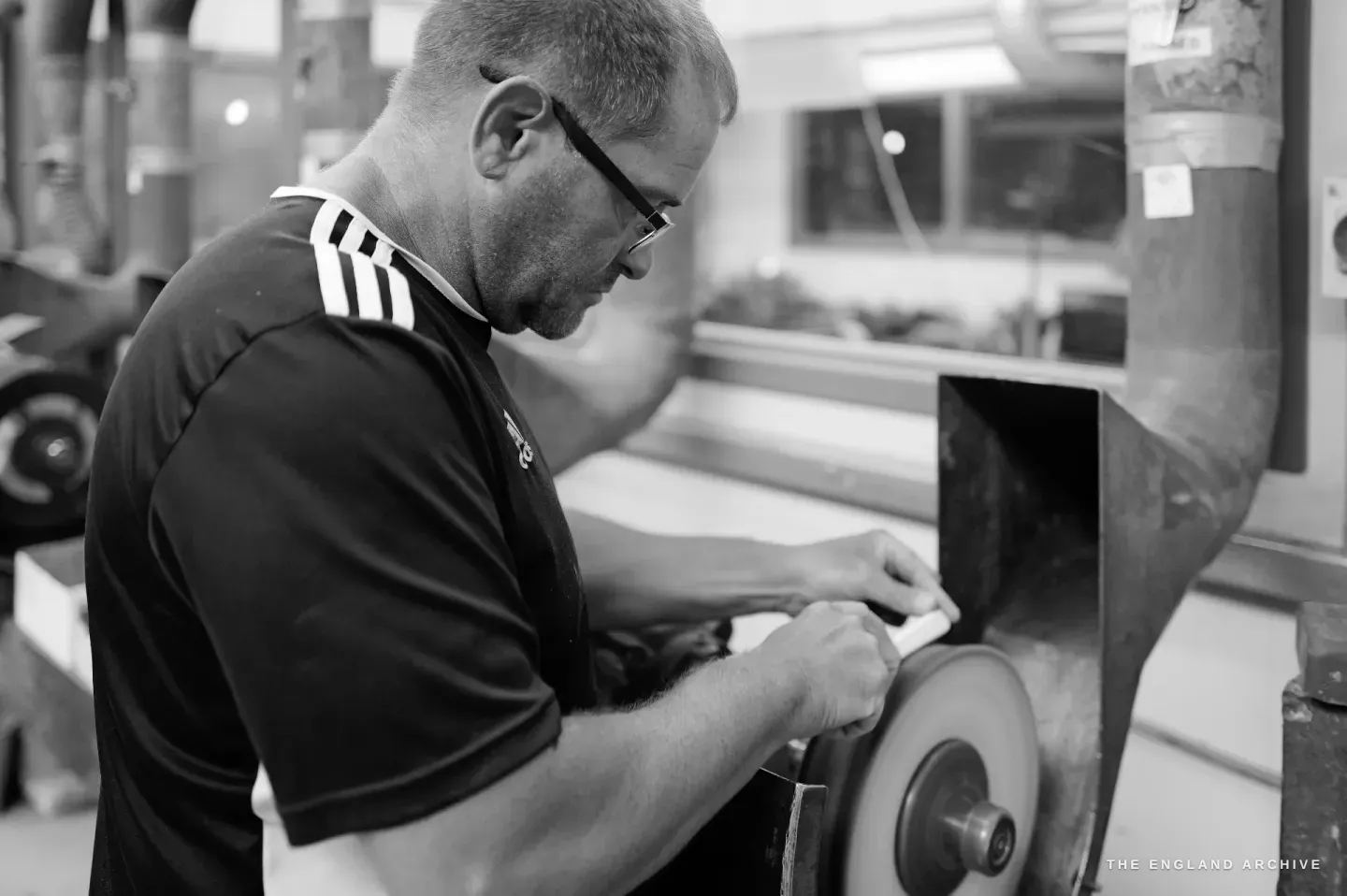 Paul in profile working a piece against a glazing wheel at the bench, a window of the workshop behind him.