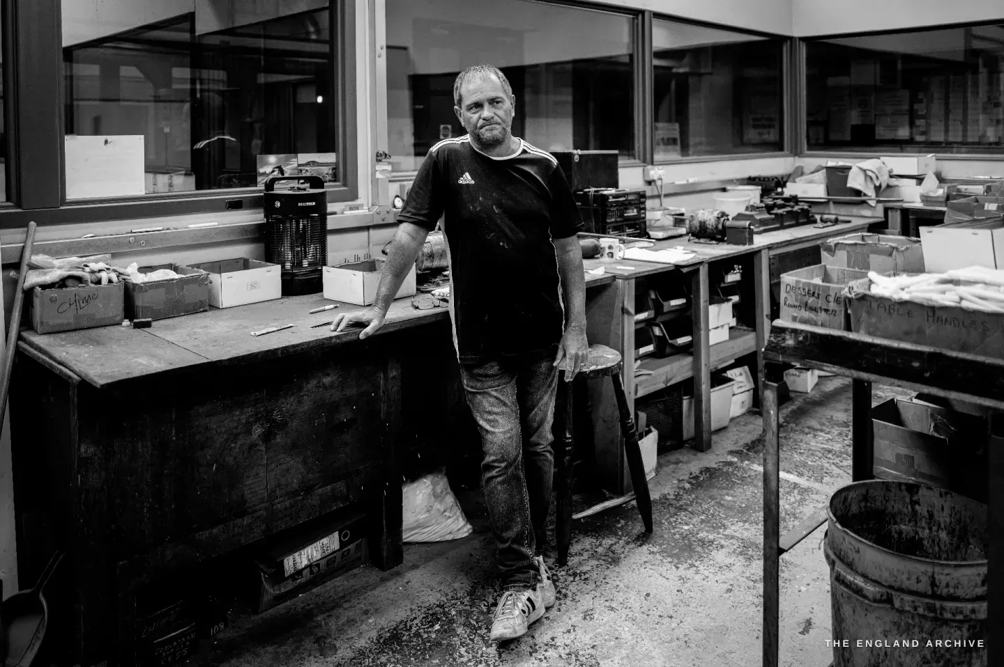 A full-length portrait of Paul standing easy against his workbench in the middle of the finishing room, benches and a galvanised bin around him.
