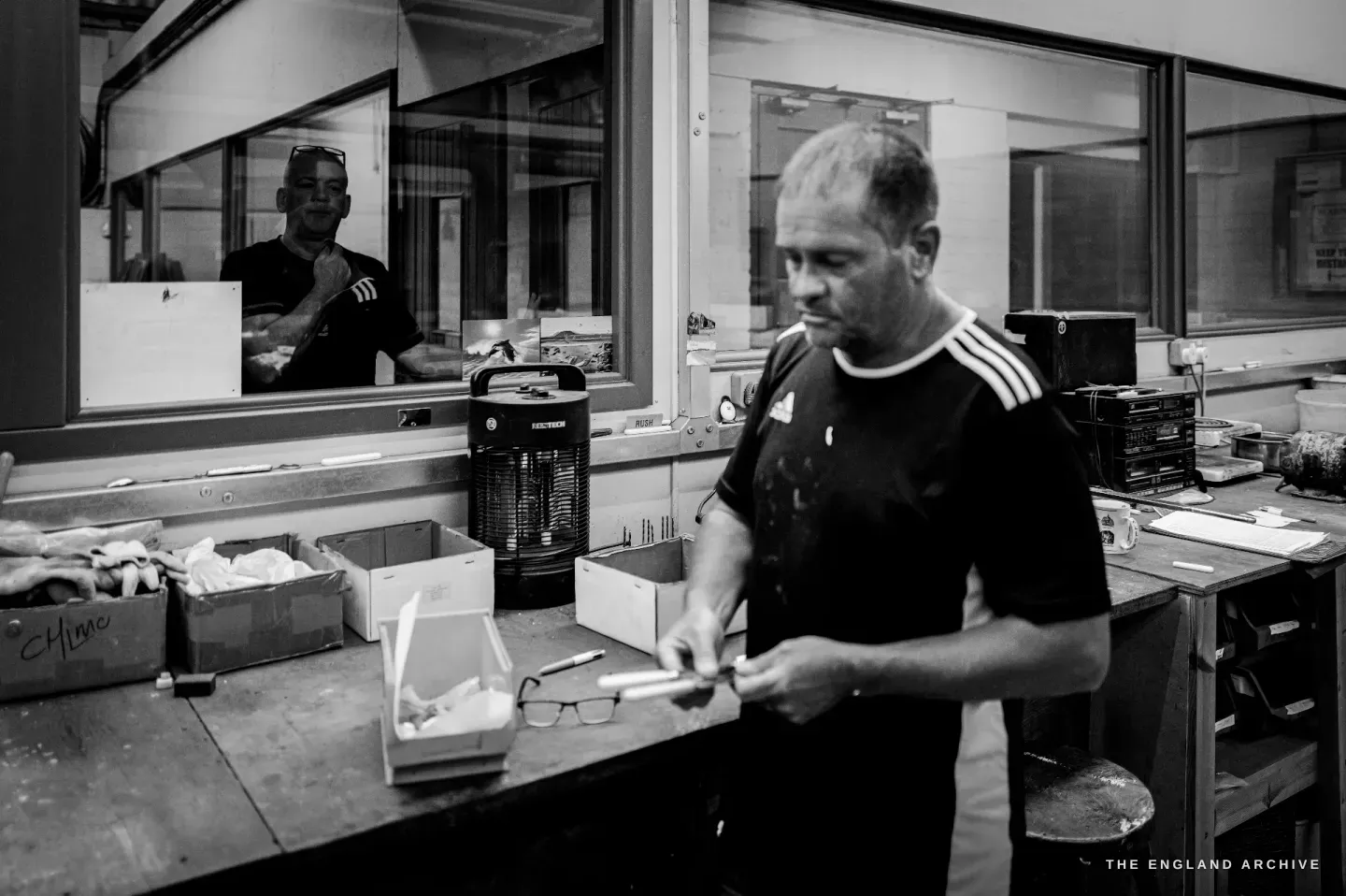 Paul at his bench handling a finished knife, his own reflection caught in the glass partition to the left.