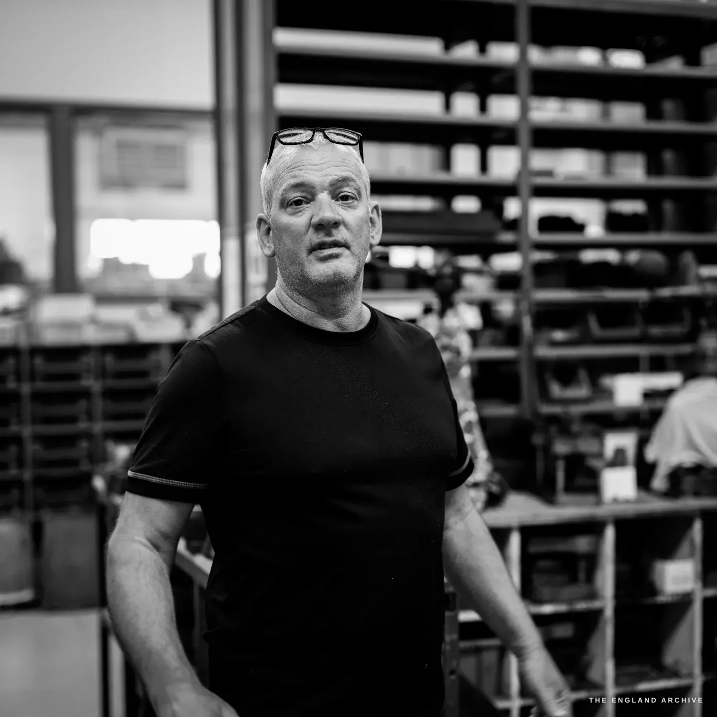Phil Benton facing the camera in the workshop, head and shoulders, mouth open mid-sentence, reading glasses pushed up on his forehead, a slatted steel rack behind him.