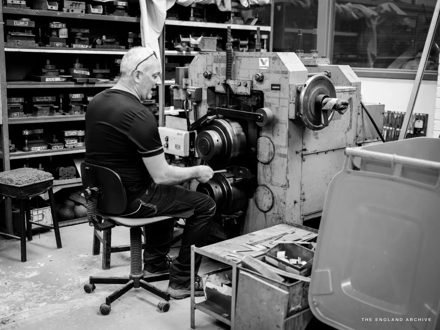 Phil seated on a sprung stool with his back to the camera, working at a horizontal rolling machine with stacked rollers, shelves of small parts boxes covering the wall in front of him.