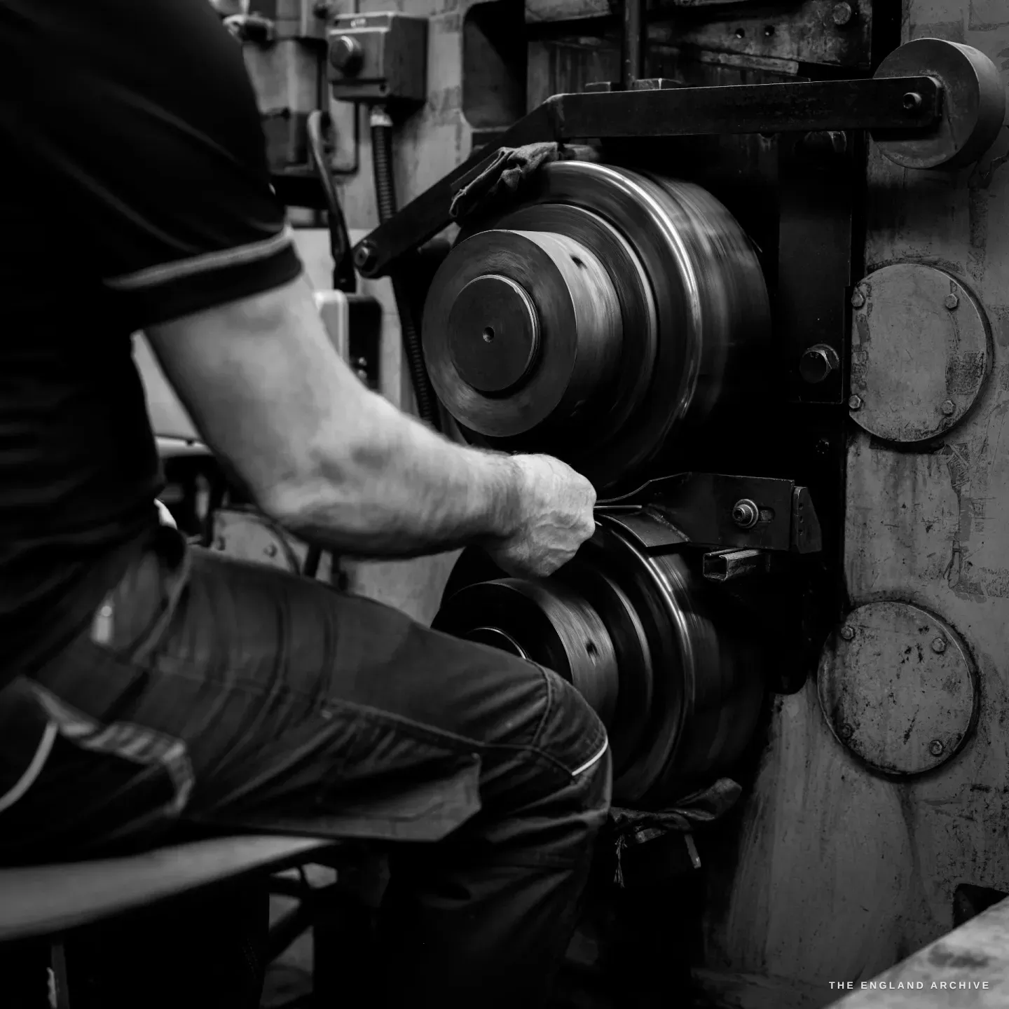 Over Phil’s shoulder: a gloved hand presses a small clamped piece against a stacked twin-wheel polishing spindle, dark wheels turning.