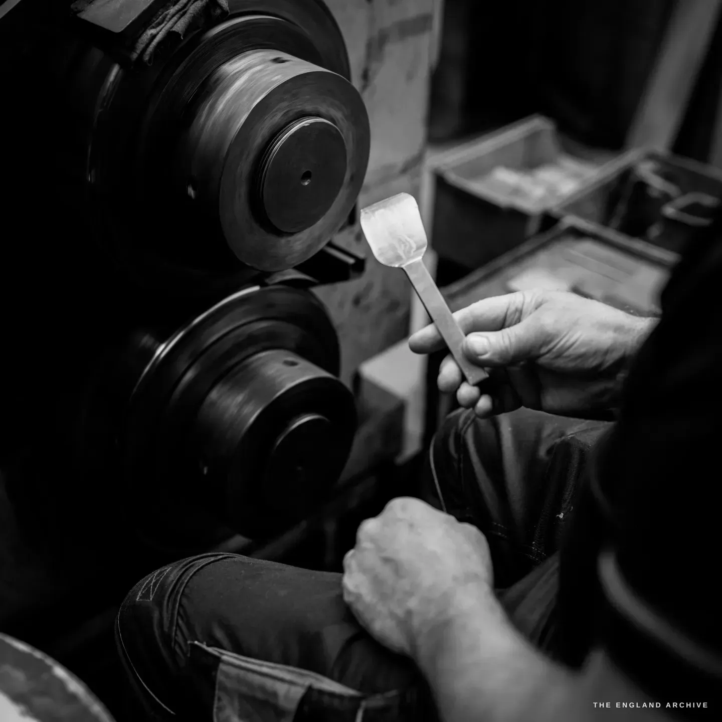 A close over-the-shoulder view of a hand holding a part-formed flat metal blank up to a stacked twin-wheel polishing spindle, the other hand braced on the knee.