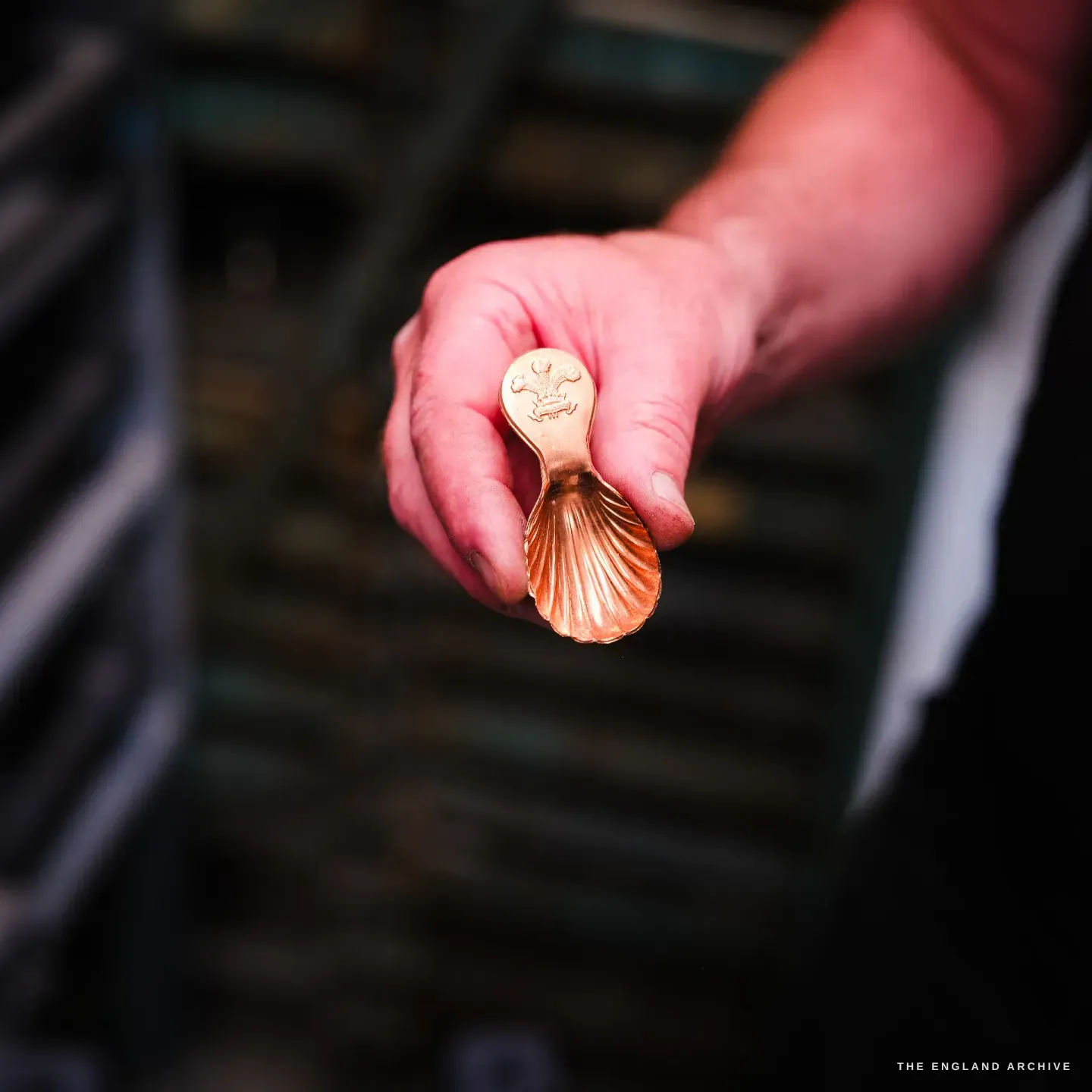 A hand holds out a small polished copper caddy spoon, its bowl fluted like a scallop shell, the stem stamped with the three-feathers Prince of Wales plumes crest.