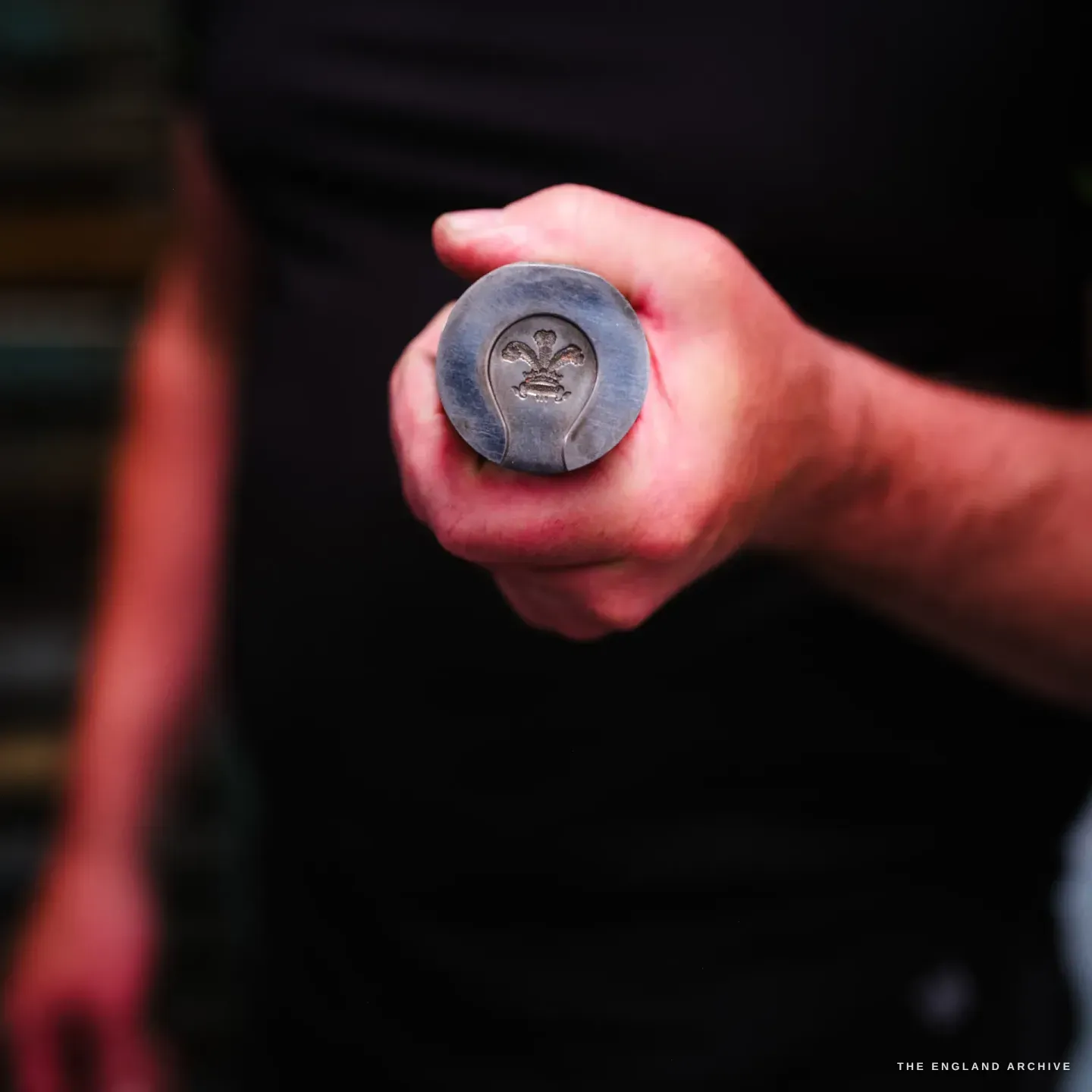 A hand grips a round steel stamping die, its face engraved with the three-feathers Prince of Wales plumes crest in a teardrop reserve.