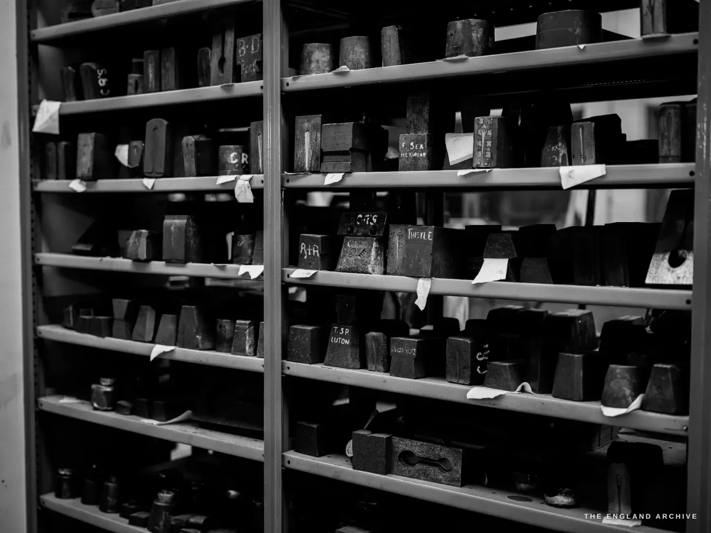 A tall steel shelving bay packed with iron die-blocks, many carrying paper labels and chalk codes.