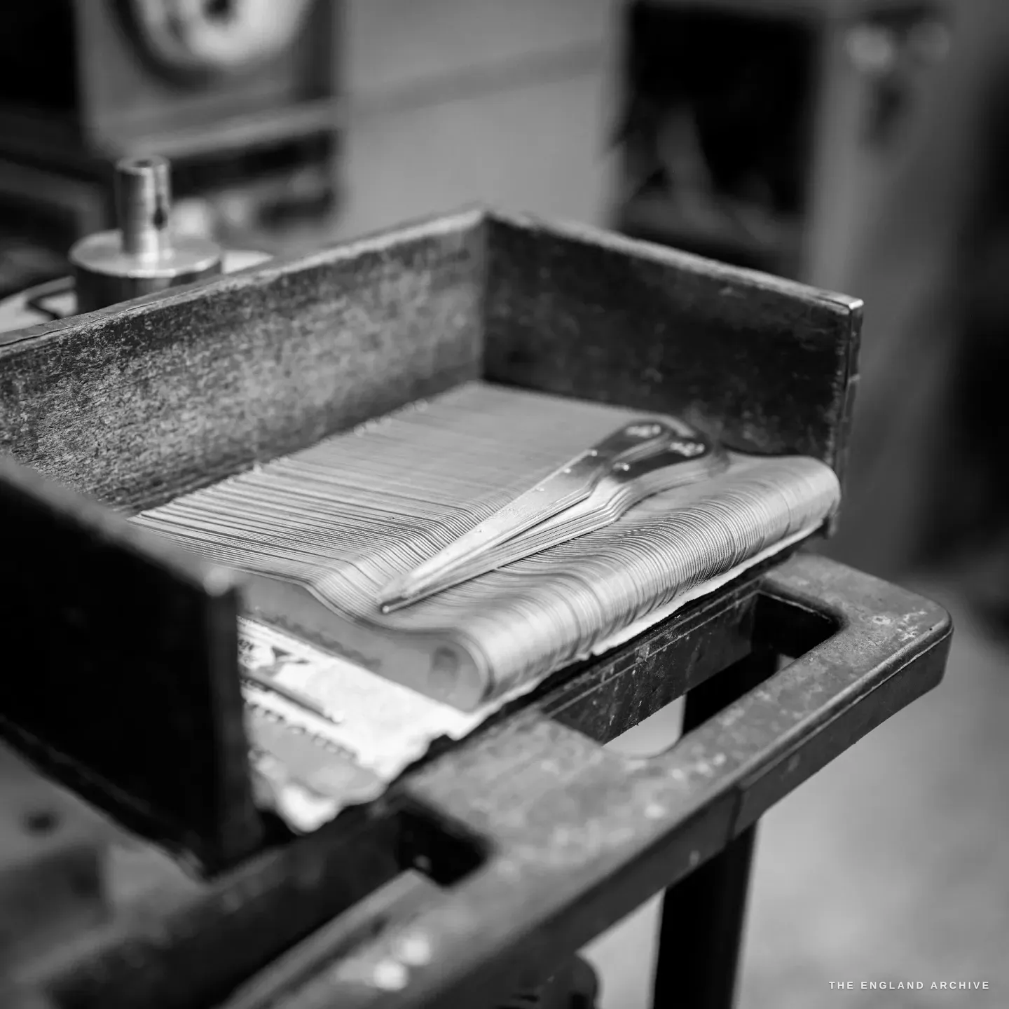 A blackened steel tray on a bench holding a fanned stack of thin cutlery blanks, one finished handle laid across the top.