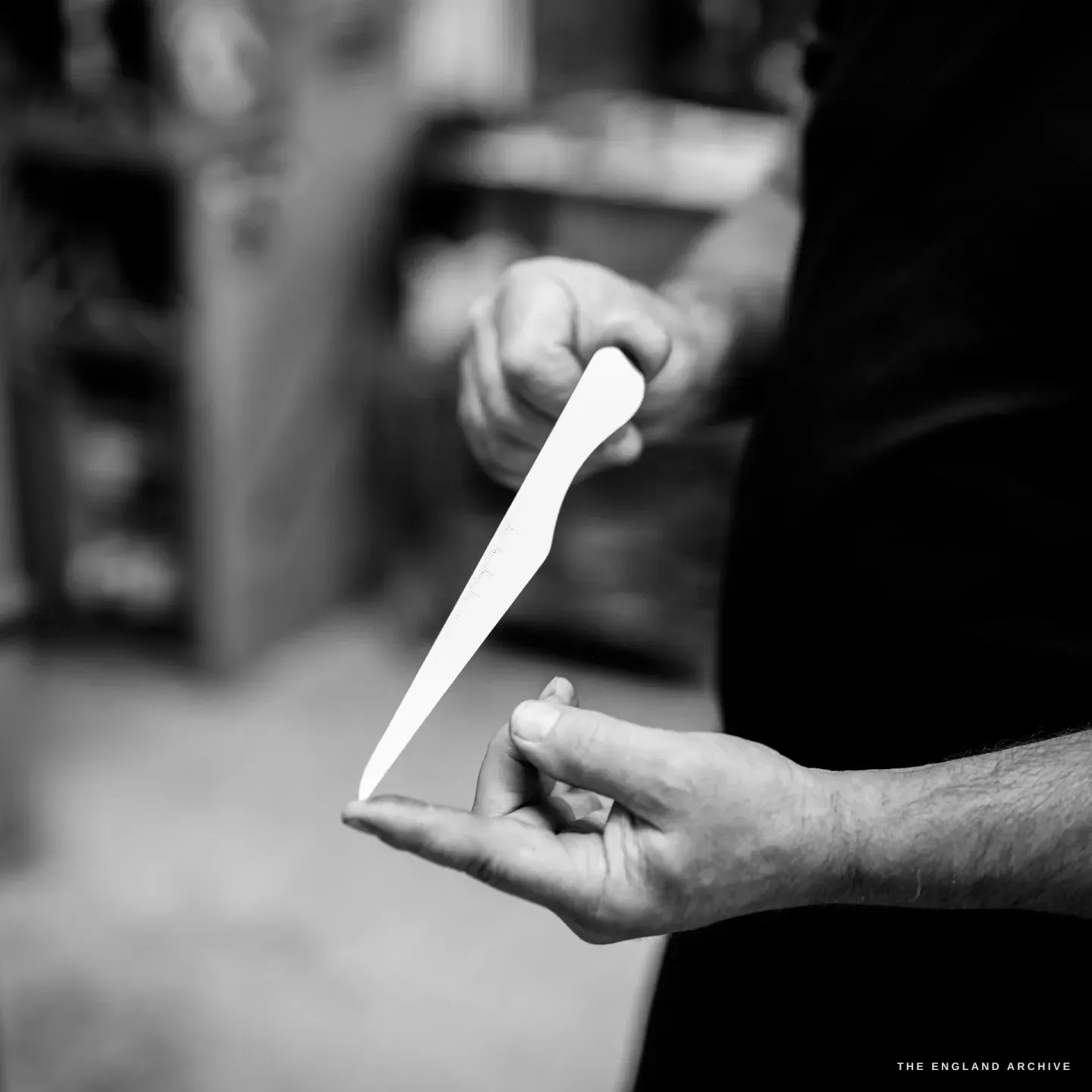 Phil holds a slim pale metal handle blank upright between finger and thumb, faint graduation marks along it, out-of-focus shelving behind.