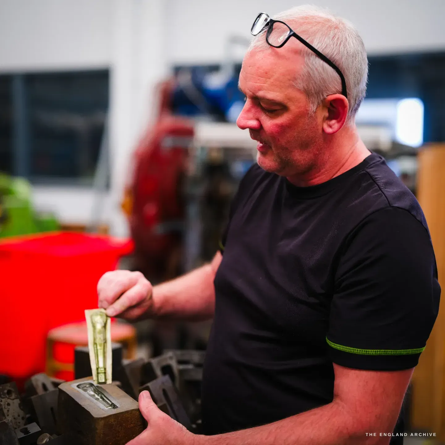 Phil in profile, glasses up on his forehead, lowering a moulded handle pattern into a rusted iron flask among rows of other mould boxes.