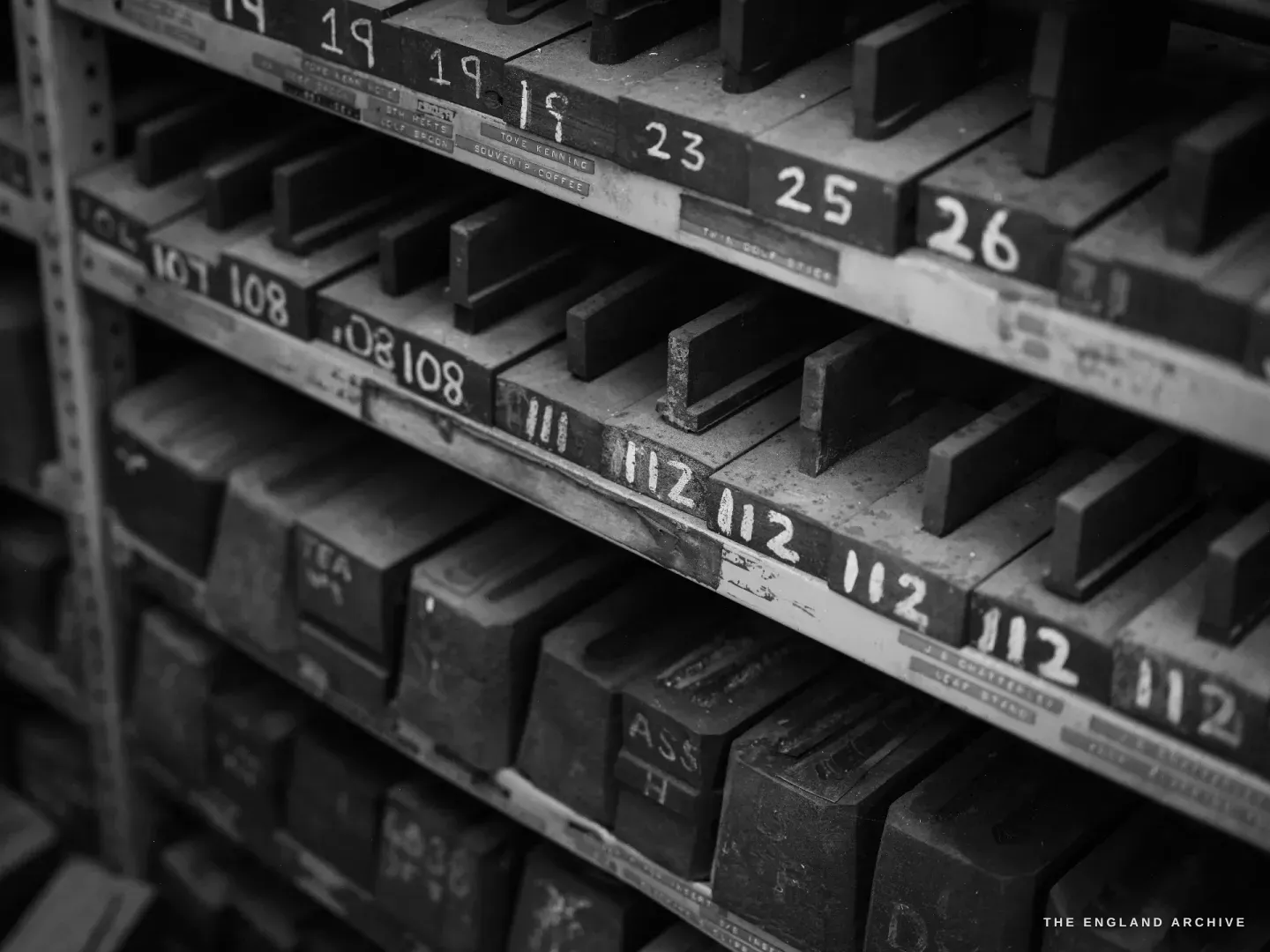 An angled view along steel die-storage shelves, the front rails chalk-numbered 19, 23, 25, 108, 112, iron stamps stacked beneath.