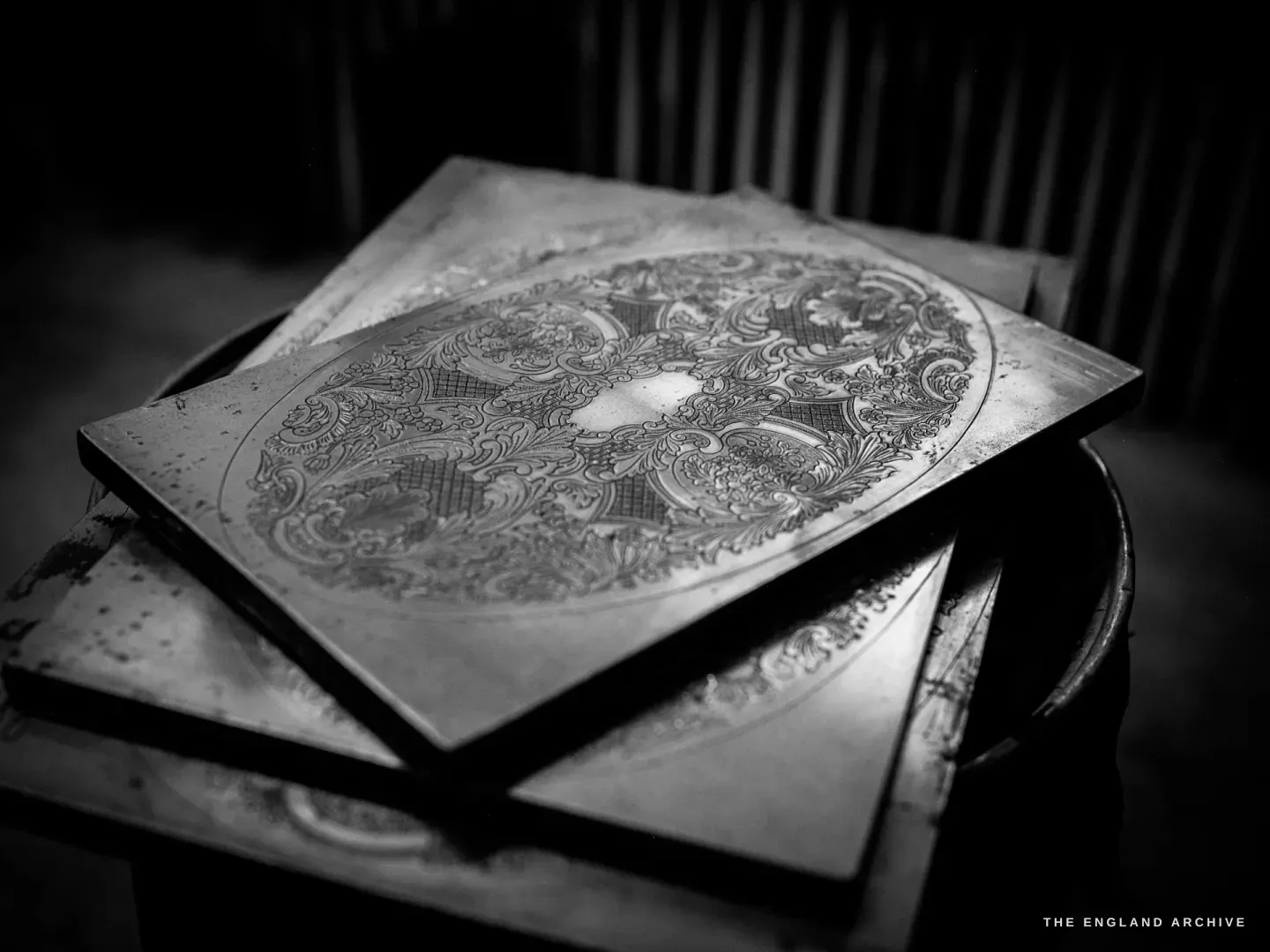 A heavy engraved steel die plate resting on a stool, its face cut with an intricate circular paisley and scroll pattern around a blank cartouche, in low light.